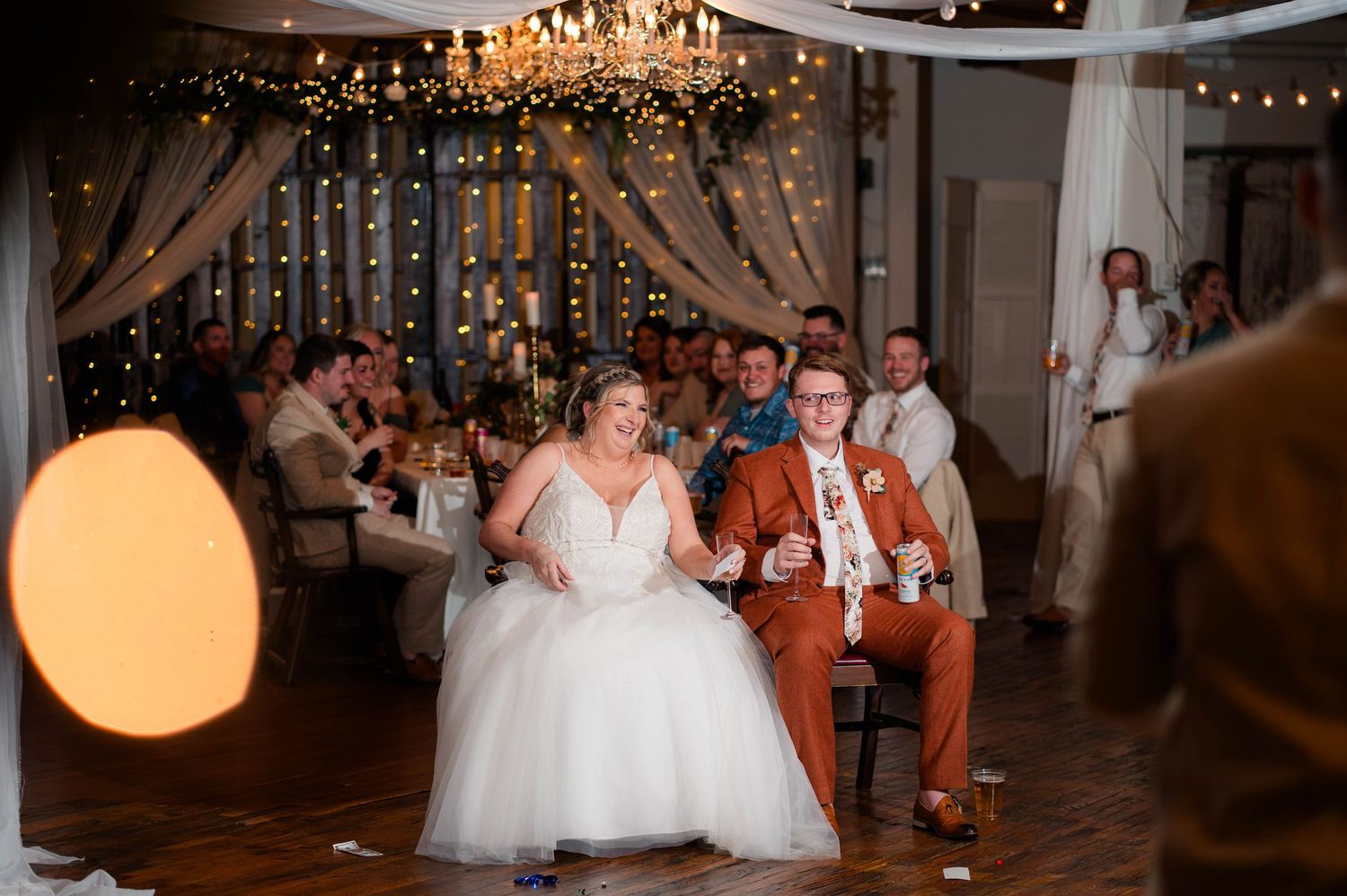 Bride and groom laugh during wedding reception game surrounded by cheering guests indoors.