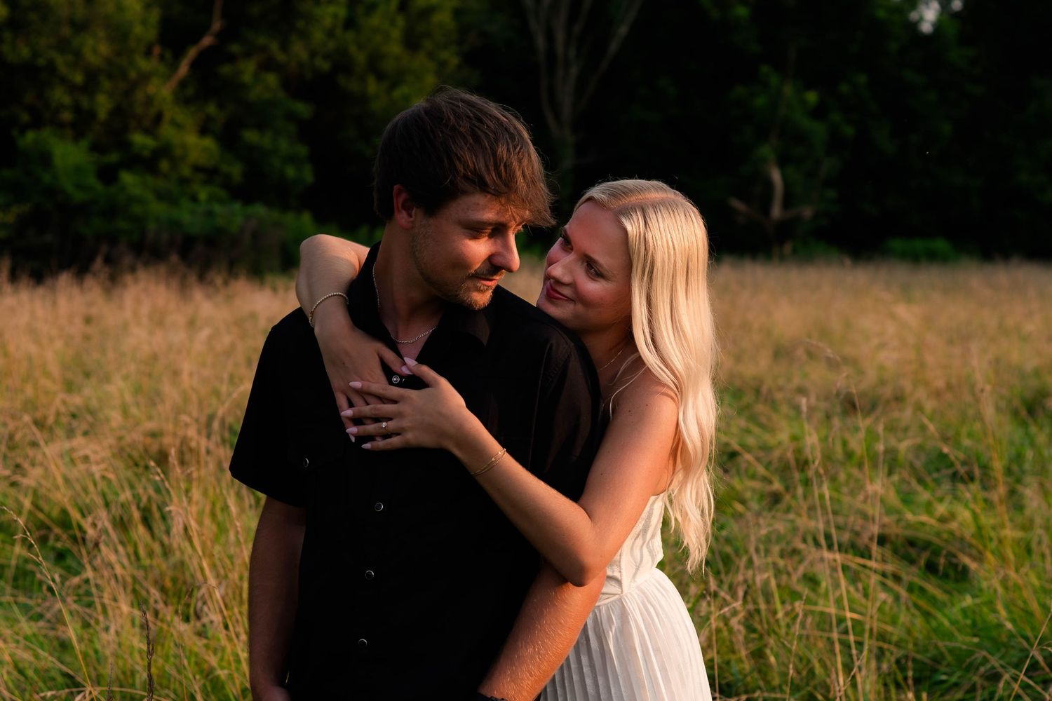 Couple embracing in golden field at sunset, woman in white dress smiling at man in black shirt.