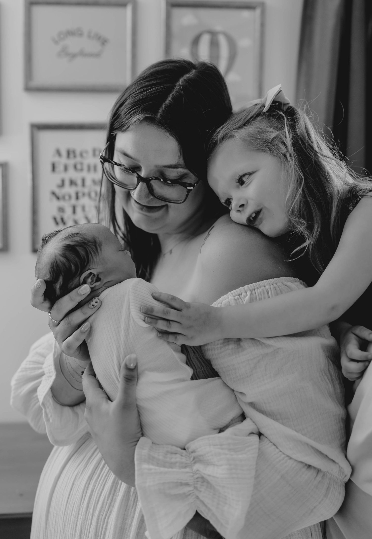 Black and white photo of mother holding newborn while toddler daughter hugs her warmly.
