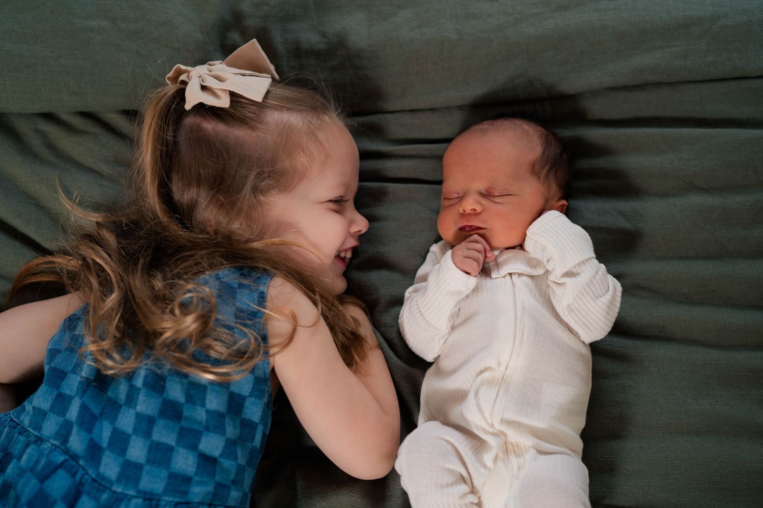 A smiling toddler girl in a blue dress lying next to a sleeping newborn baby on a green surface.