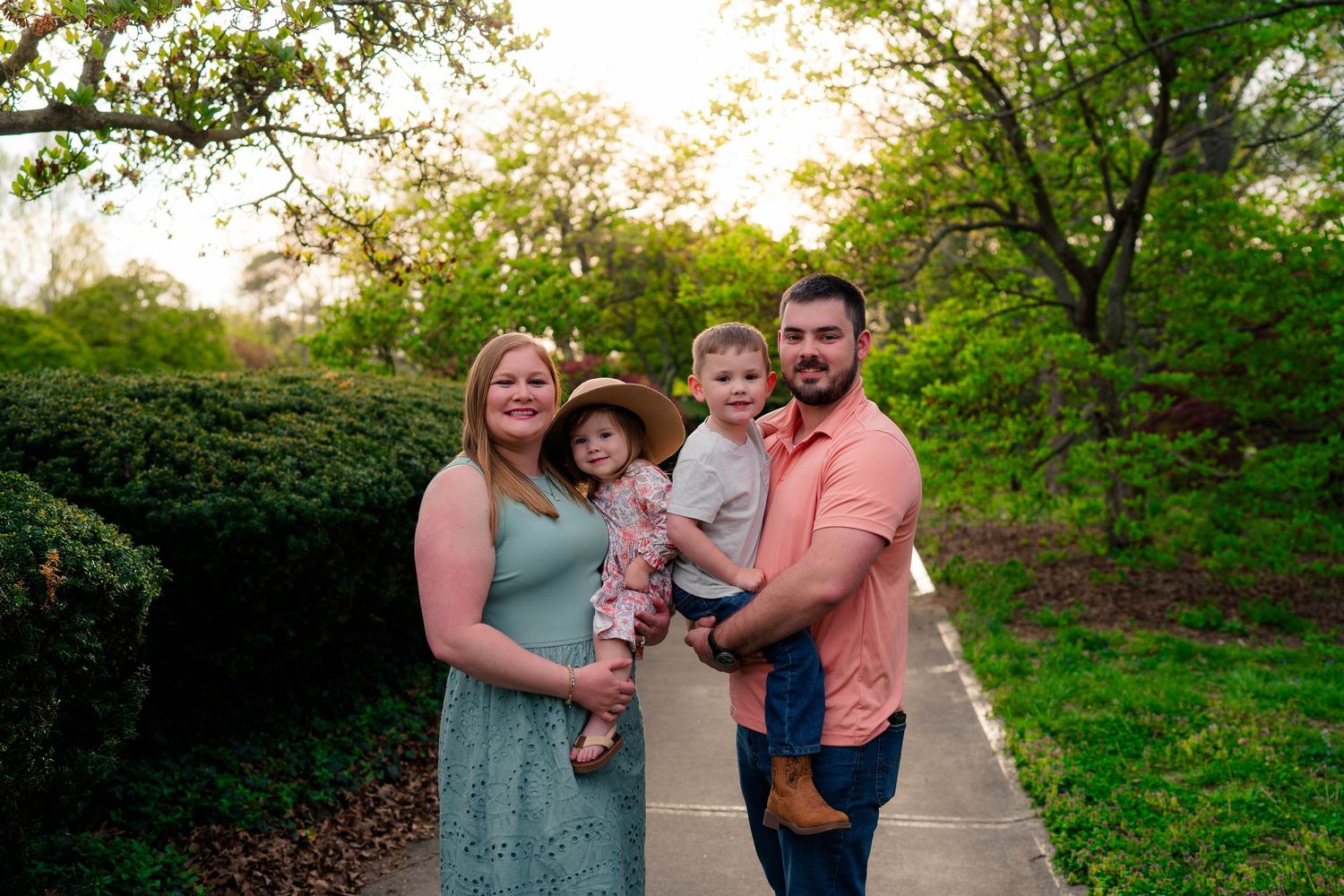 A happy family of four poses on a garden path at sunset, parents holding two young children in spring attire.