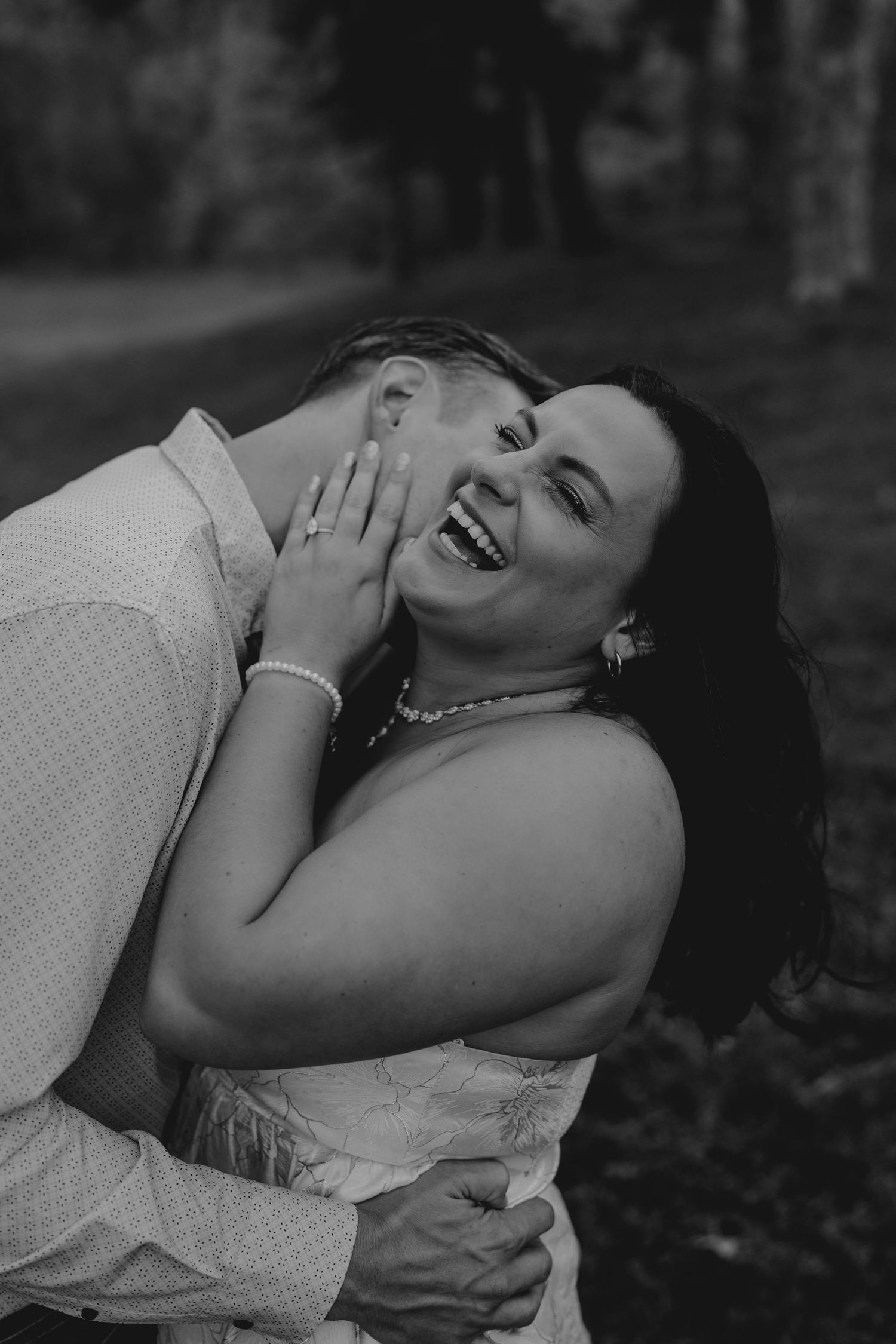 Black and white photo of a laughing woman being kissed on the cheek by her partner during engagement photos.