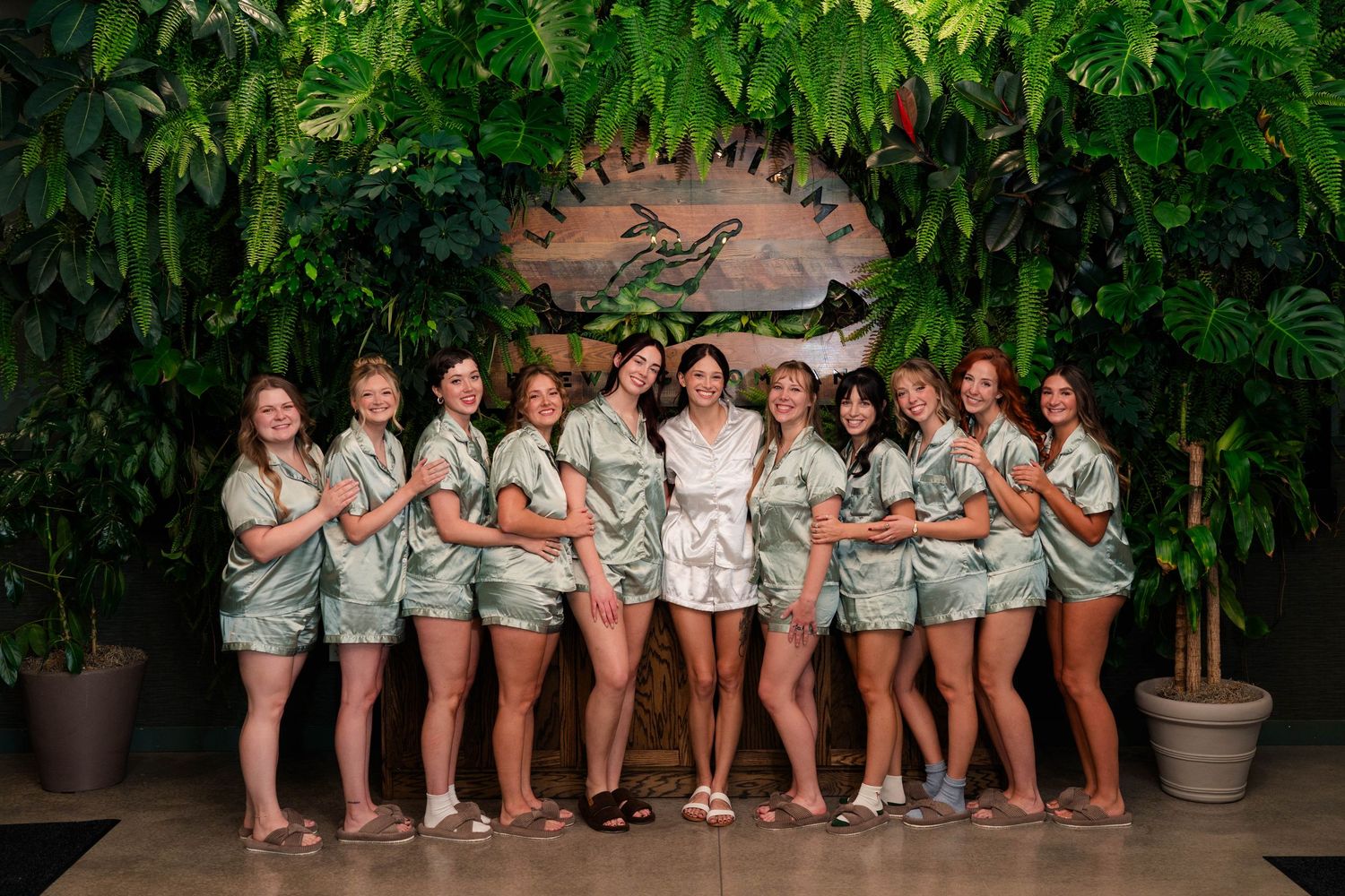 Bridal party in matching sage green satin pajamas posing together in front of a tropical green wall backdrop.