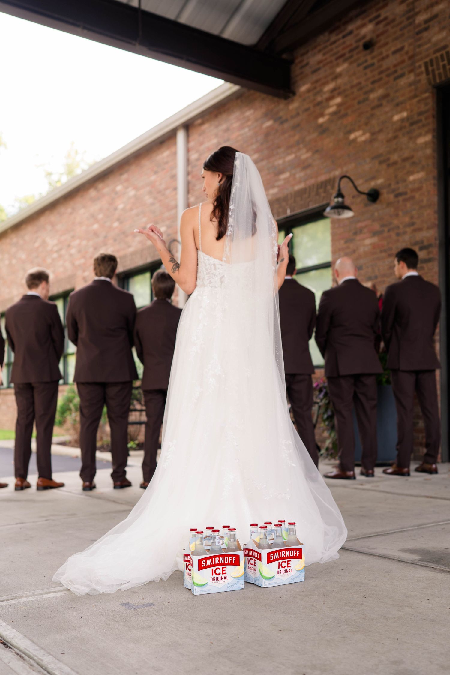 Bride in white gown surprises groomsmen with Smirnoff Ice bottles at wedding venue.