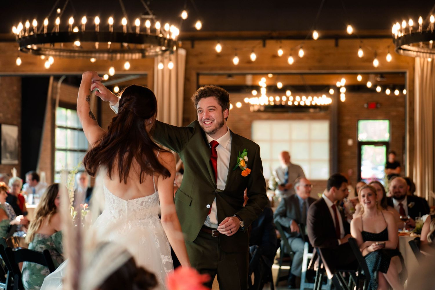 Smiling groom in olive suit twirls bride during first dance at romantic wedding reception.