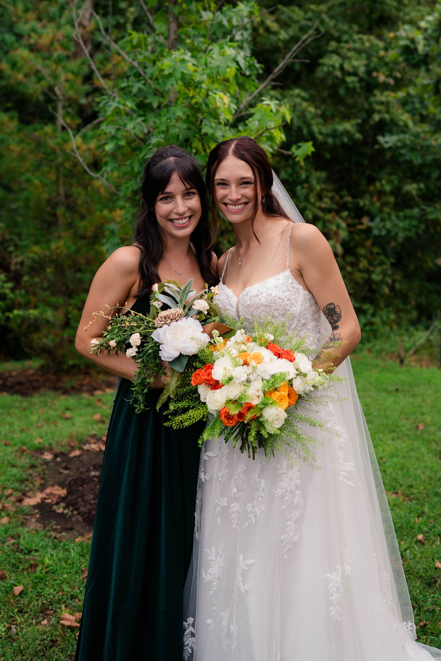 Bride in lace gown and bridesmaid in green dress smile holding colorful floral bouquets outdoors.