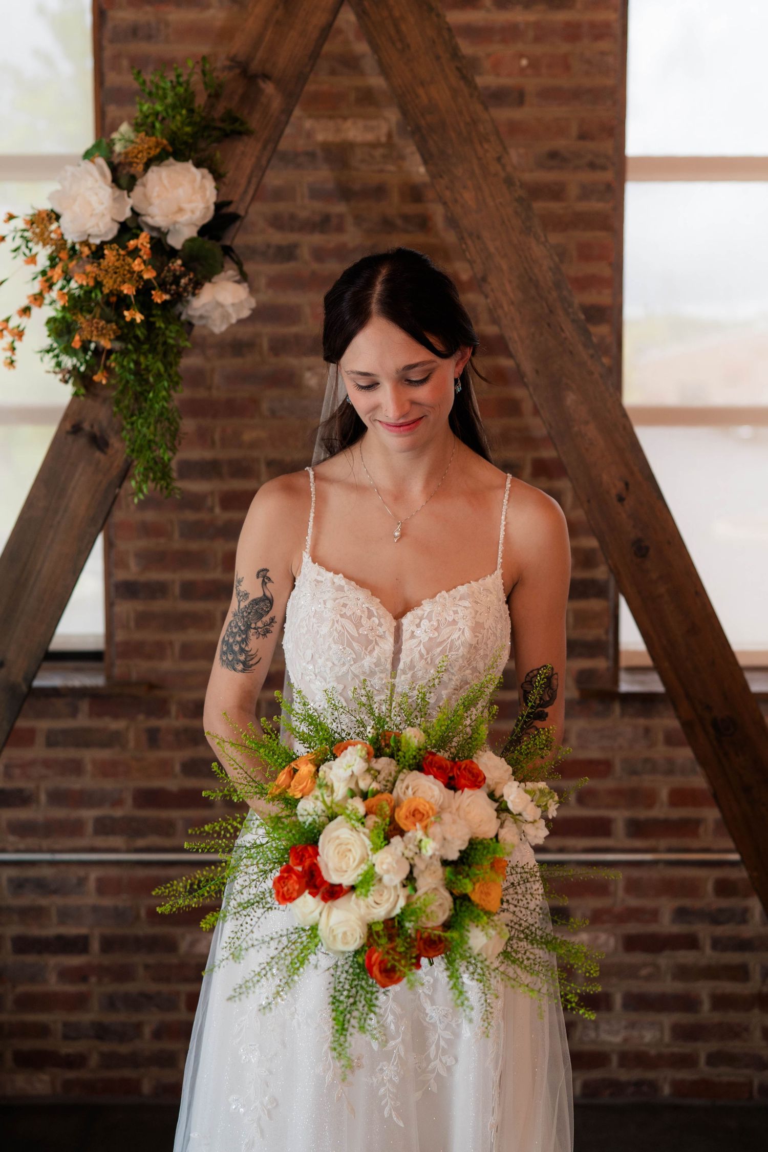 Radiant bride in lace gown holds vibrant orange and white bouquet before rustic wooden arch altar.