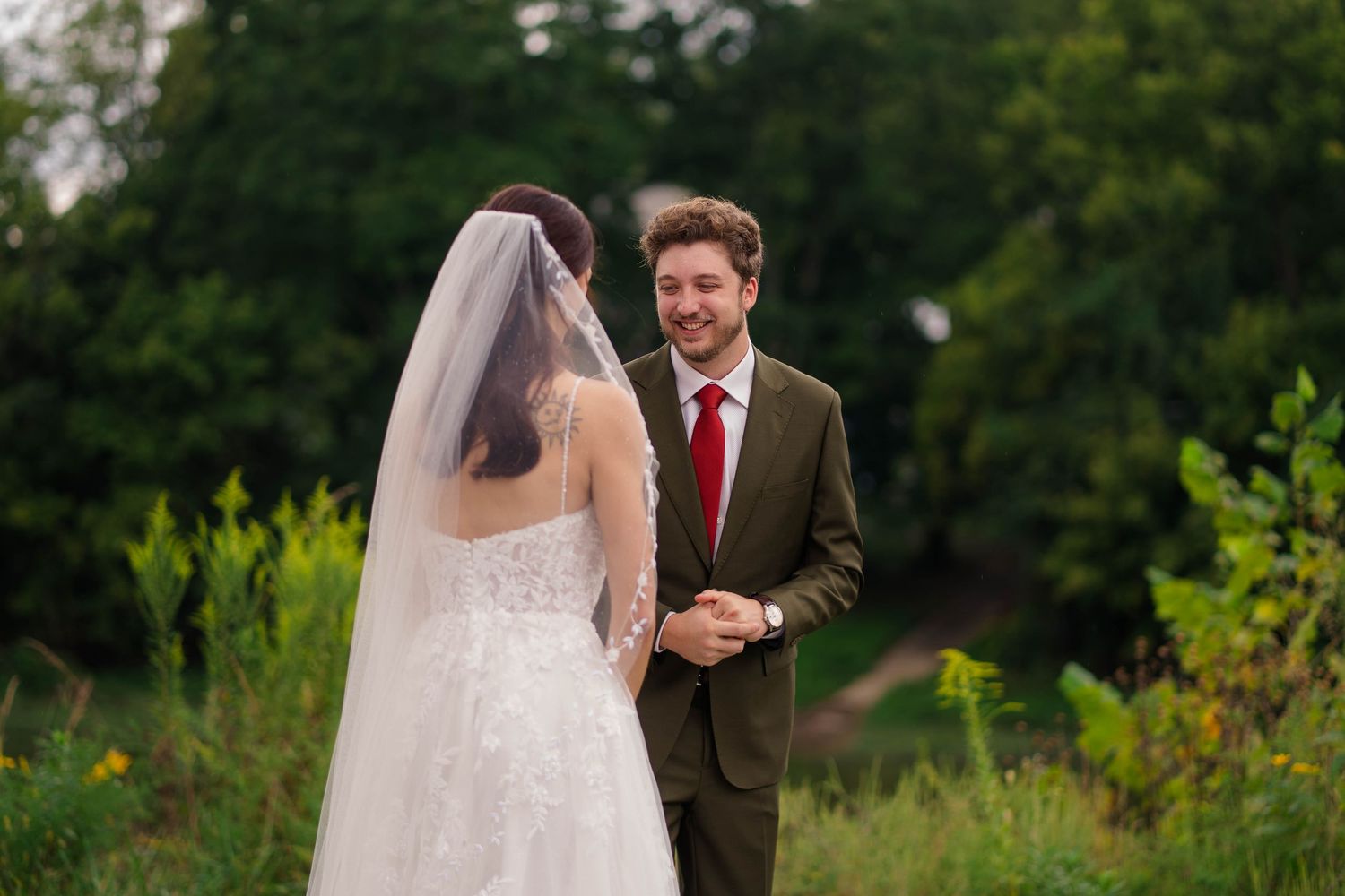 Groom in olive suit smiling at bride during outdoor first look surrounded by green foliage.