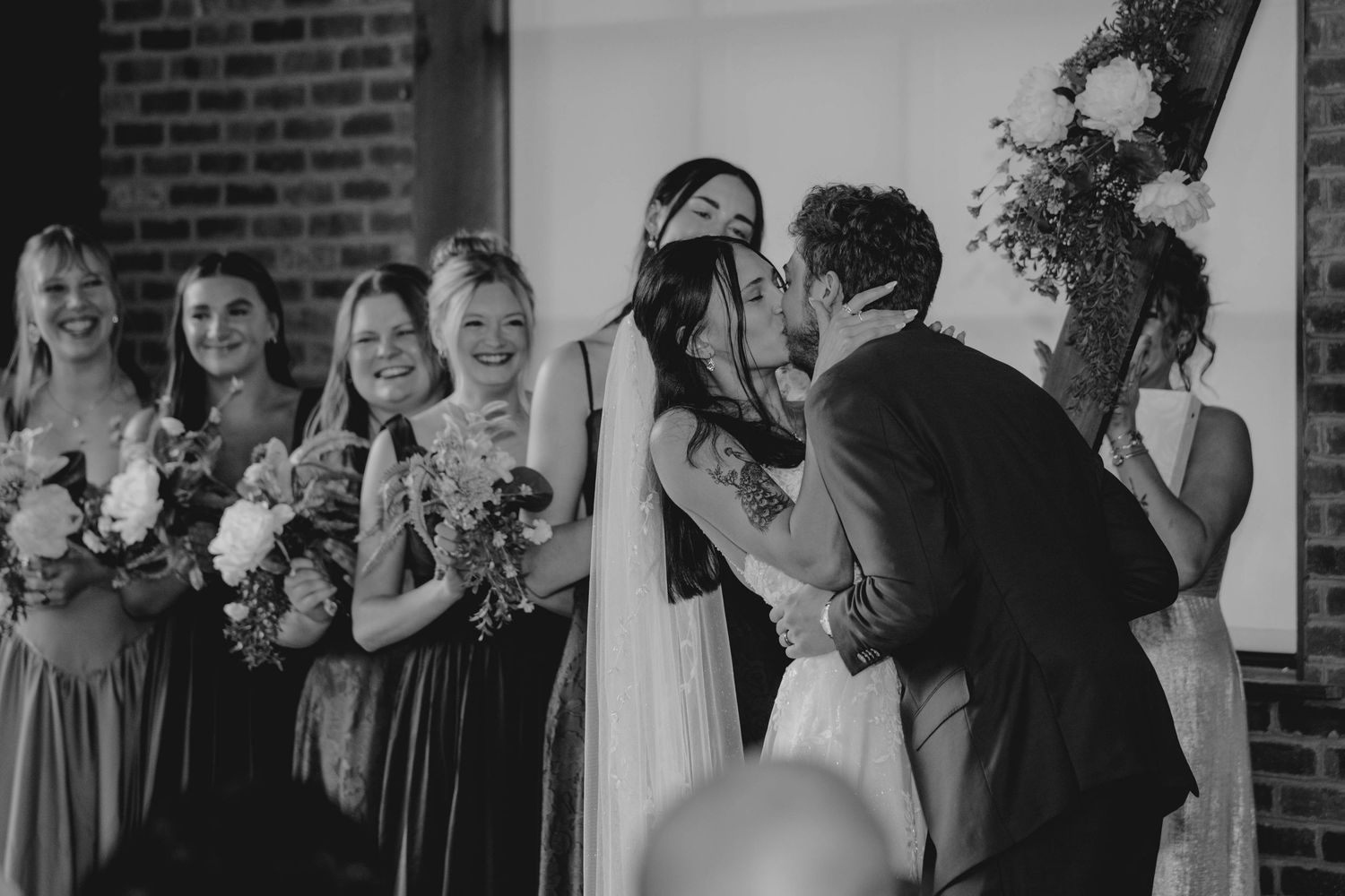 Bride and groom share first kiss surrounded by smiling bridesmaids holding floral bouquets at wedding ceremony.