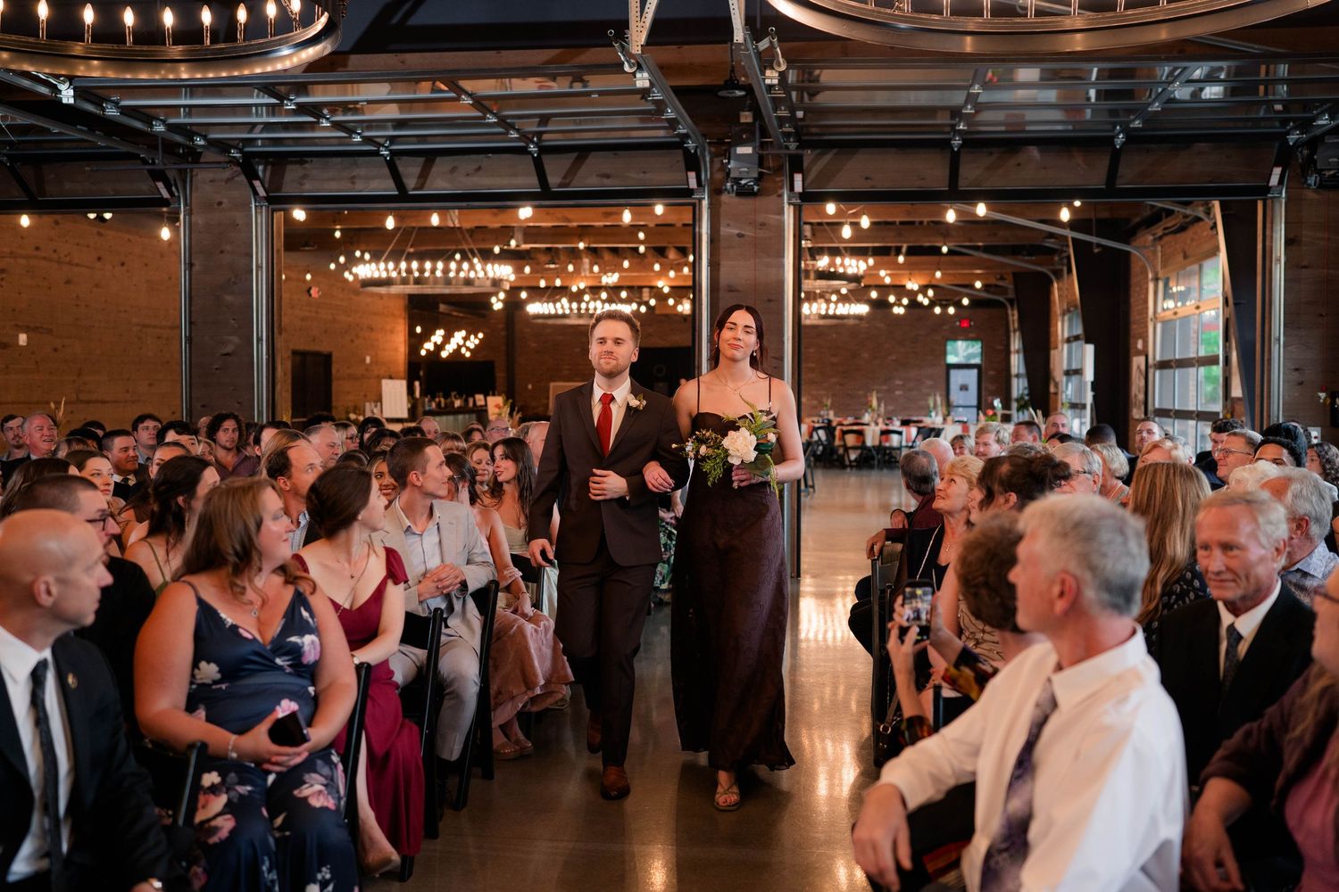 Bride walks down the aisle with escort at elegant indoor wedding venue with string lights.