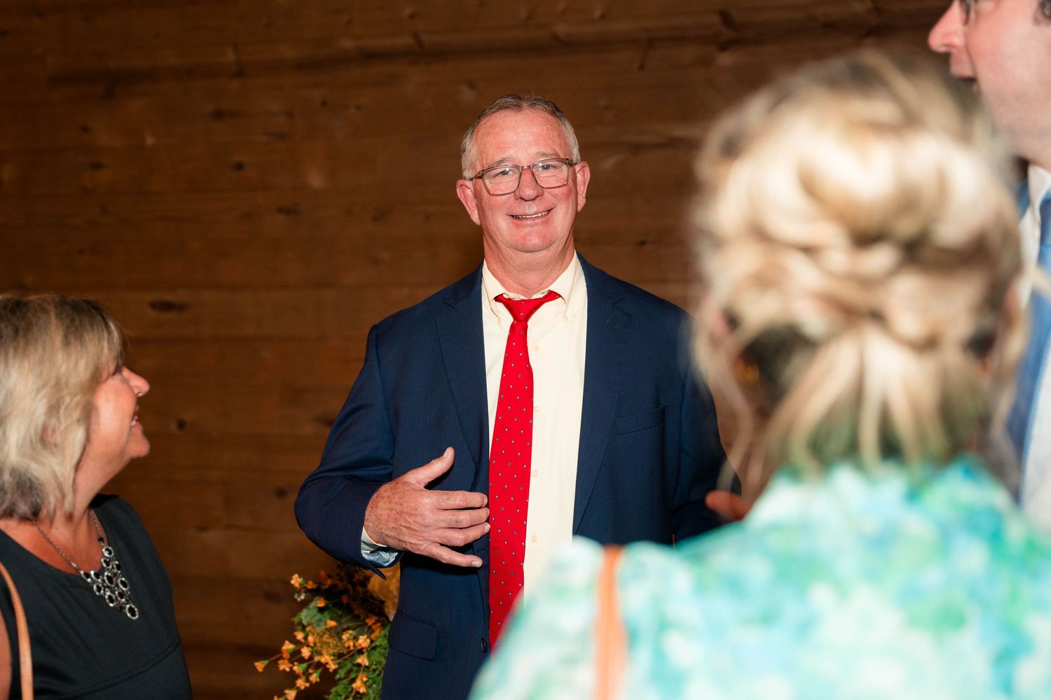 Older gentleman in navy suit and red tie smiles warmly while chatting with wedding guests.
