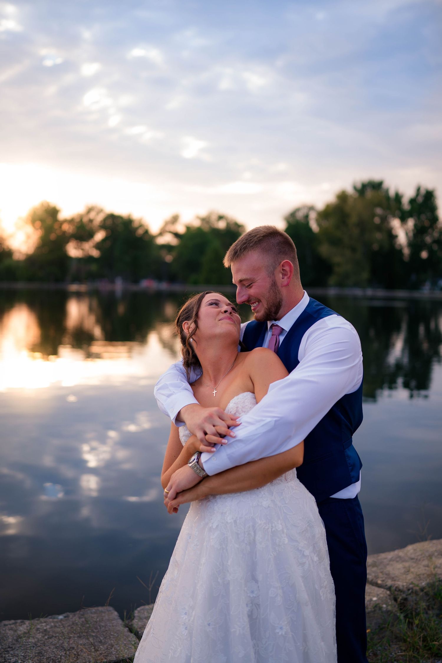 Bride and groom embrace romantically by a reflective lake at sunset, groom wrapping arms around bride in white lace gown.