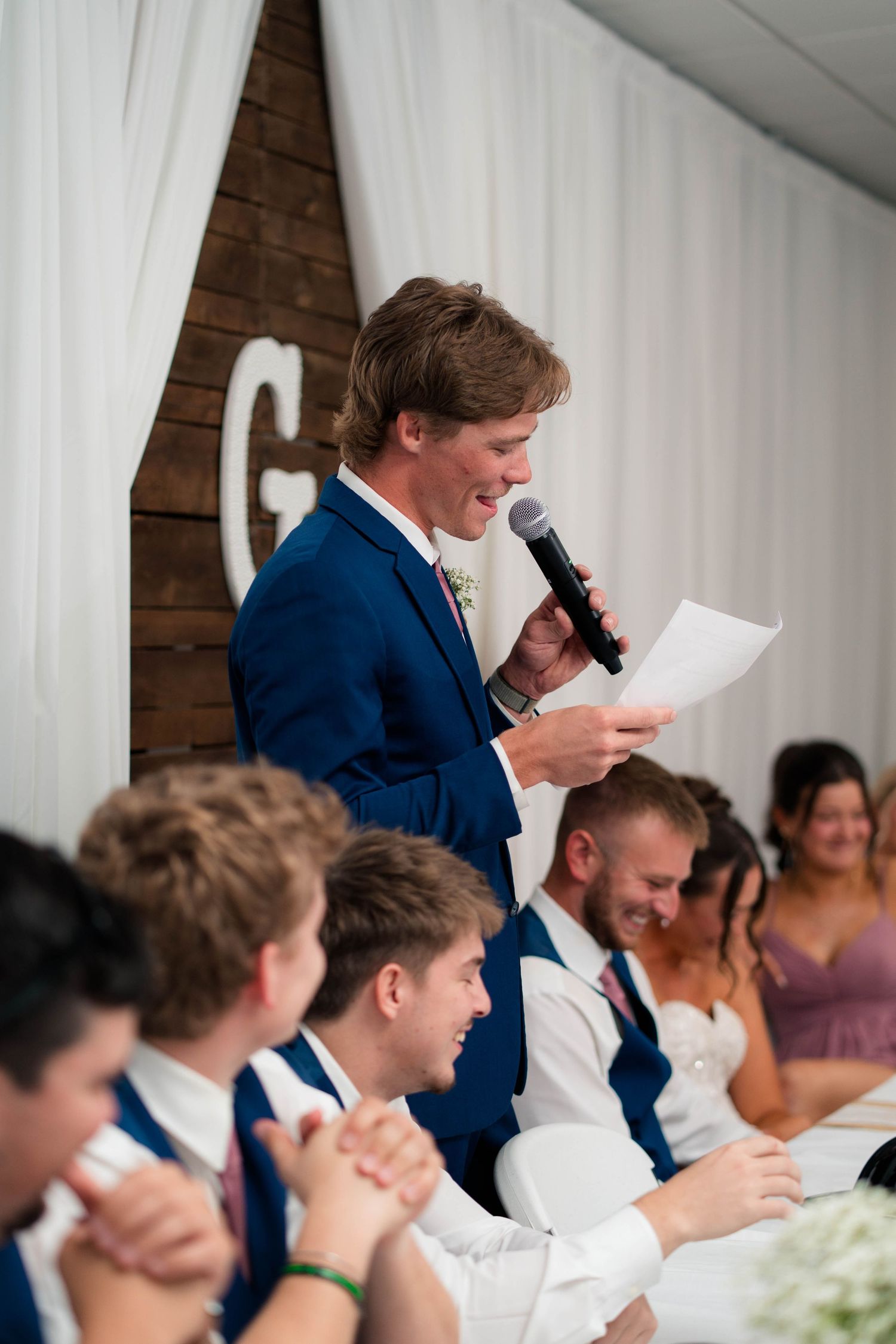 Smiling groomsman reads wedding speech into microphone while guests and bridal party laugh at reception.