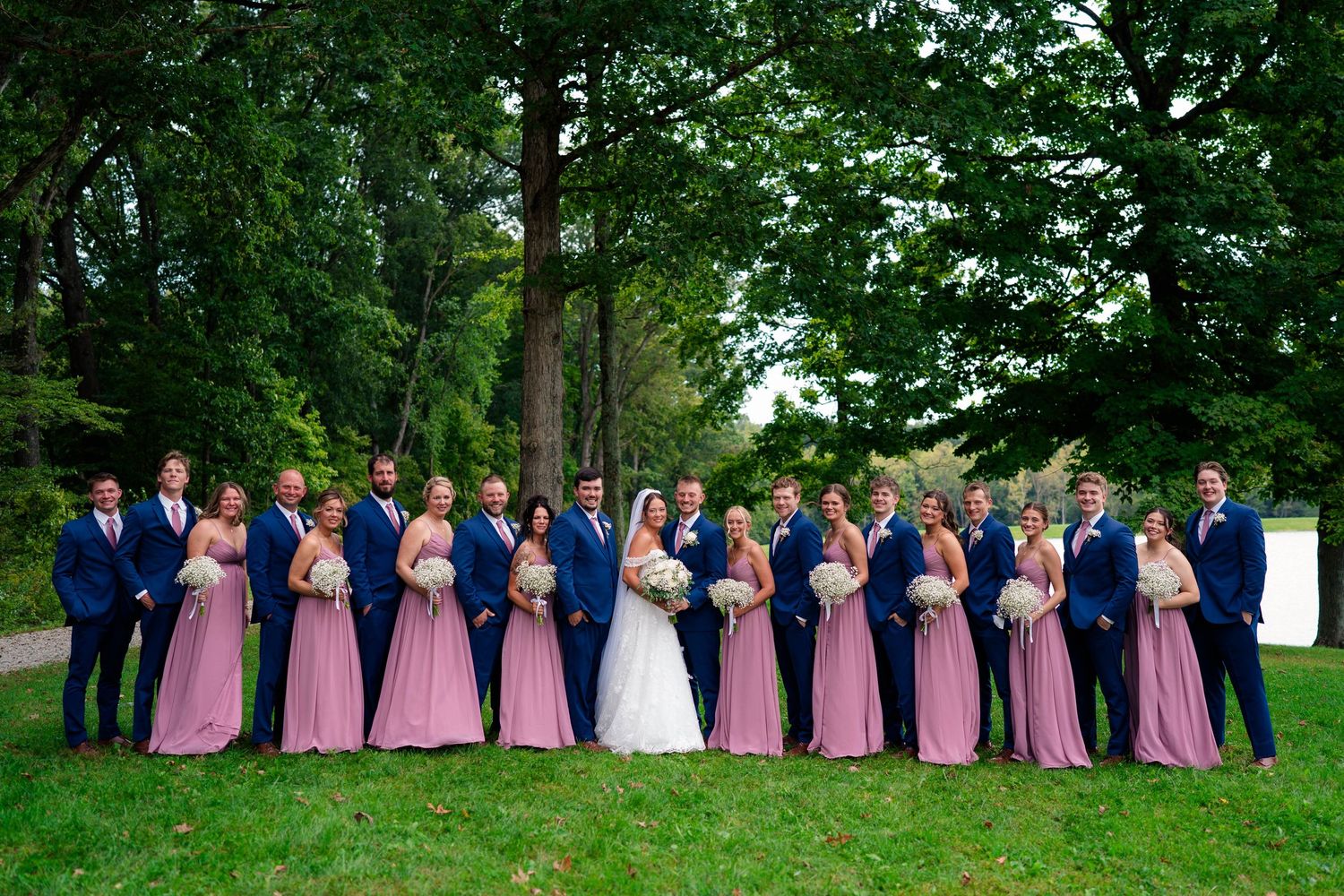 Wedding party poses together with bridesmaids in mauve gowns and groomsmen in navy suits outdoors.