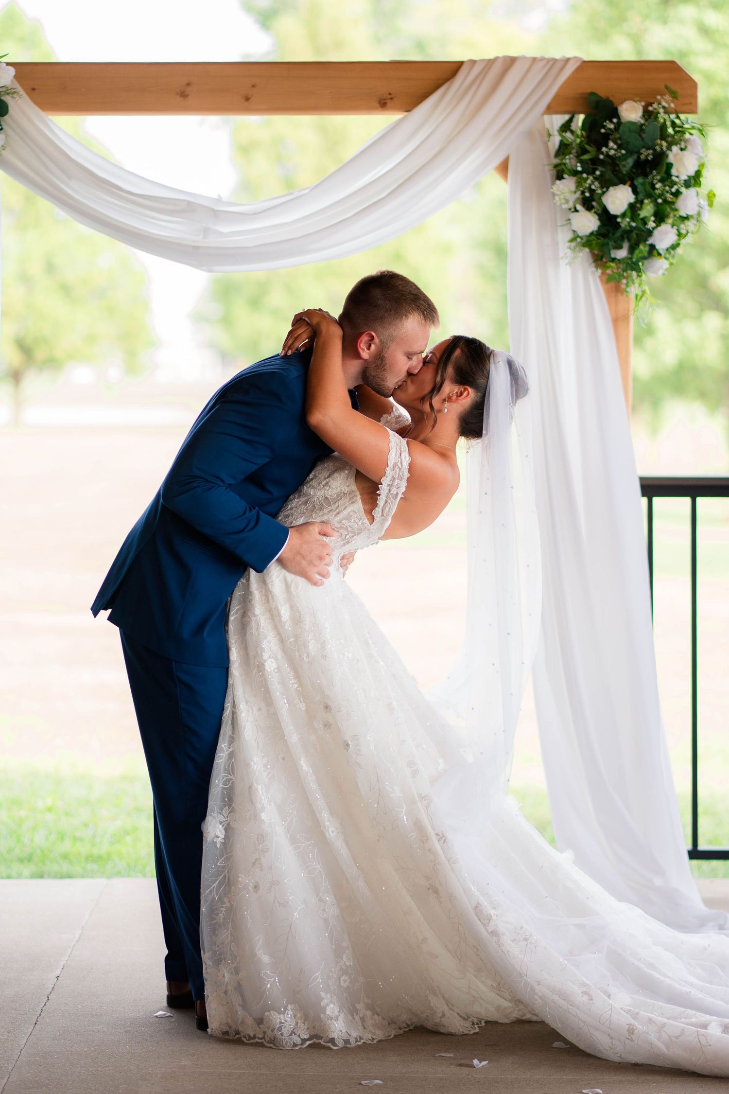 Bride and groom sharing their first kiss under a white draped arch with greenery at wedding.