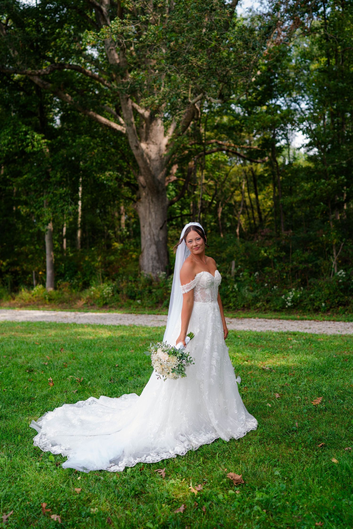 Full-length bridal portrait of bride in off-shoulder lace gown with long train, holding bouquet in green park.