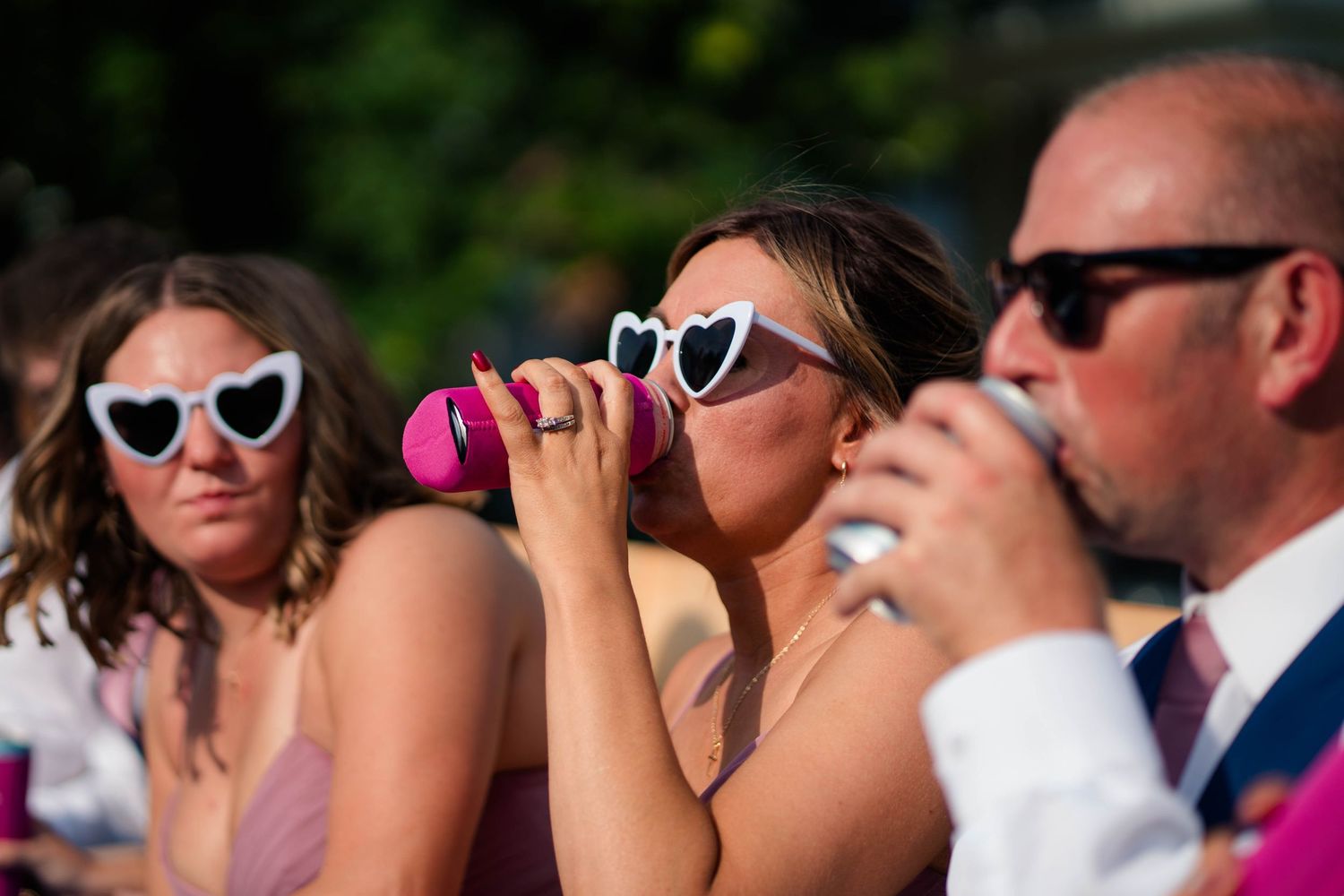 Wedding guest in heart-shaped white sunglasses drinks from pink cup alongside others at outdoor summer wedding.