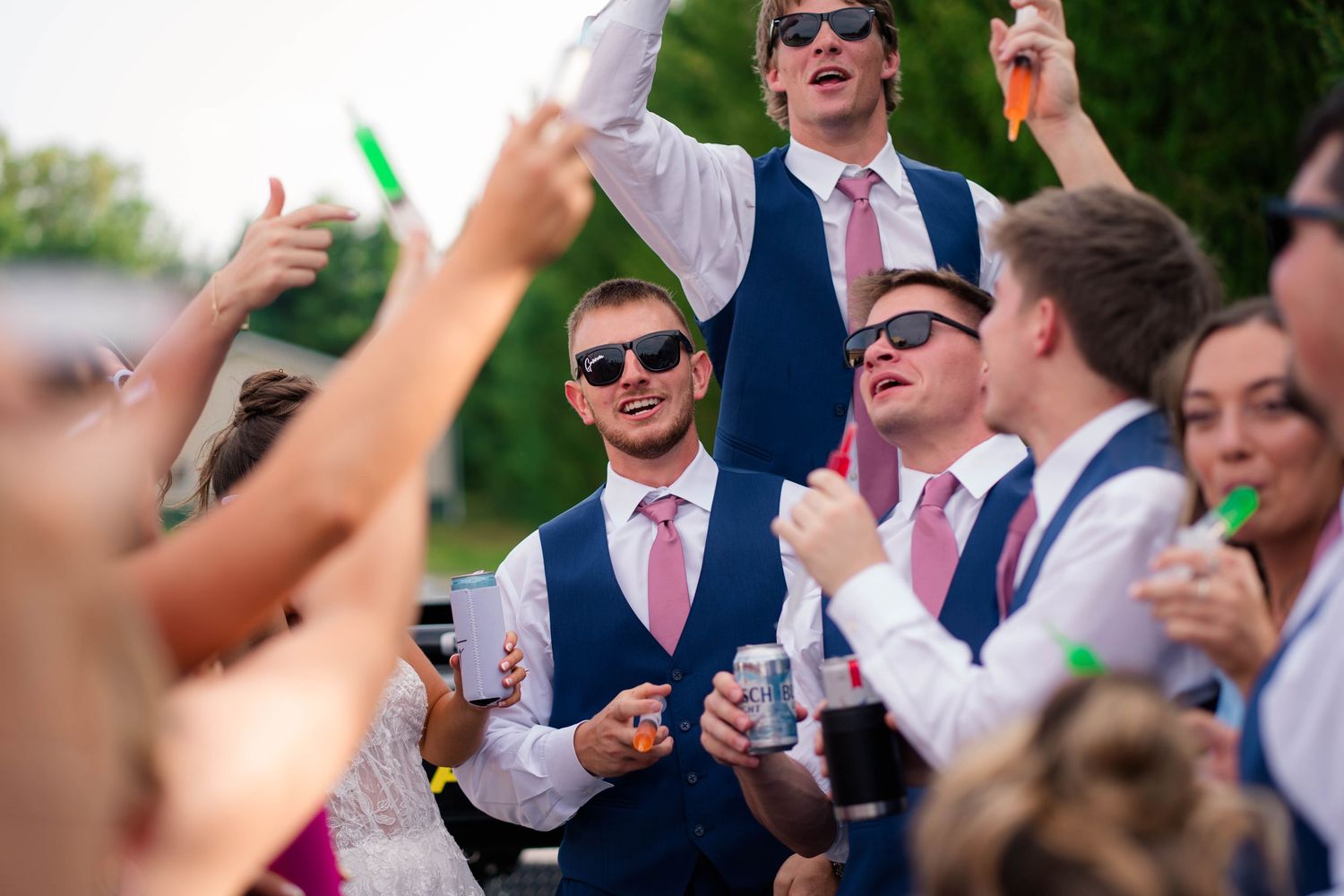 Groomsmen in navy vests and pink ties celebrating outdoors with drinks at wedding reception.