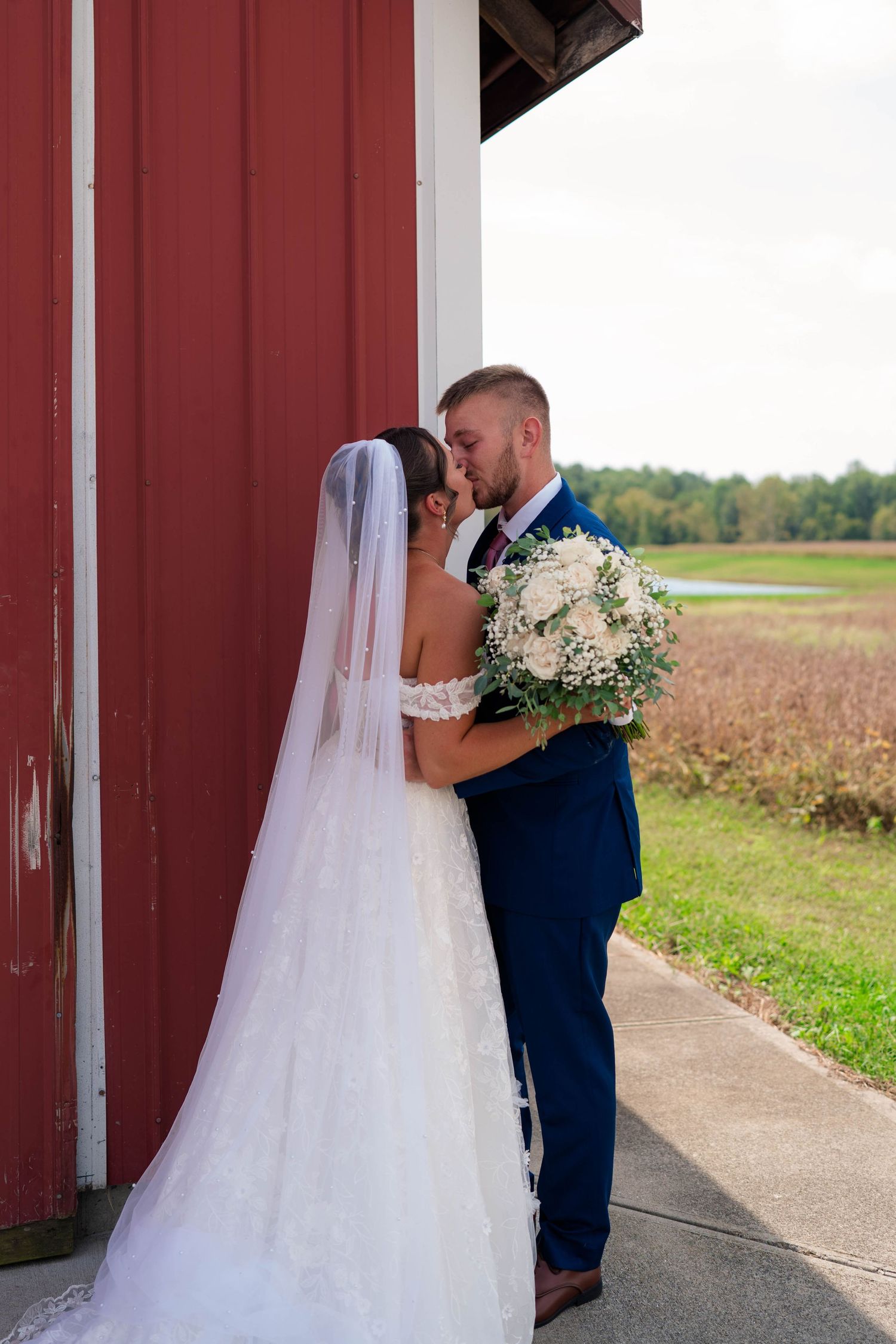 Bride and groom share a romantic kiss beside a red barn with scenic countryside views on their wedding day.