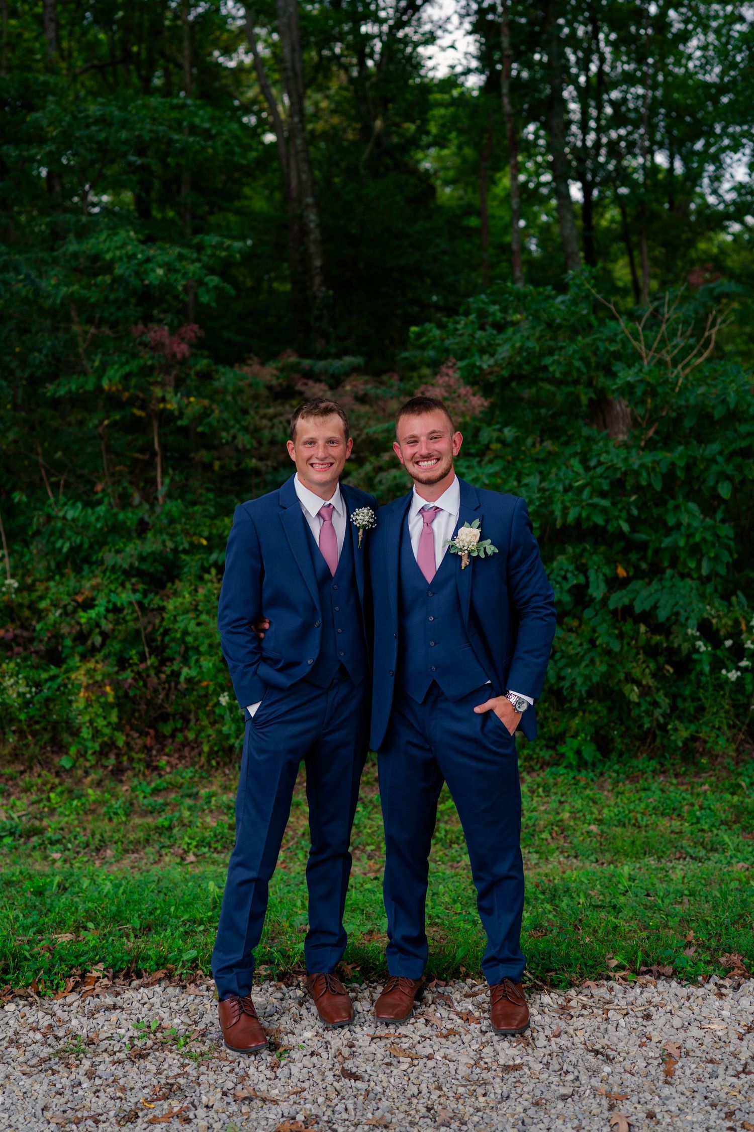 Two smiling groomsmen in matching navy three-piece suits with pink ties posing on gravel path outdoors.