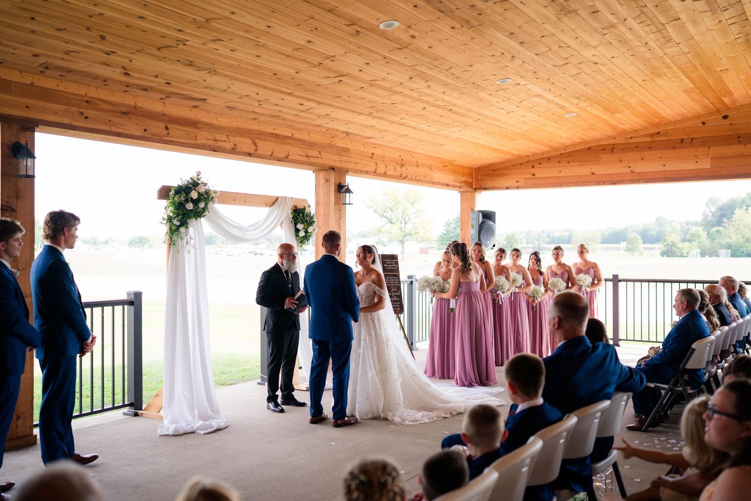 Wedding ceremony under wooden pavilion with bridesmaids in pink dresses and floral arch.