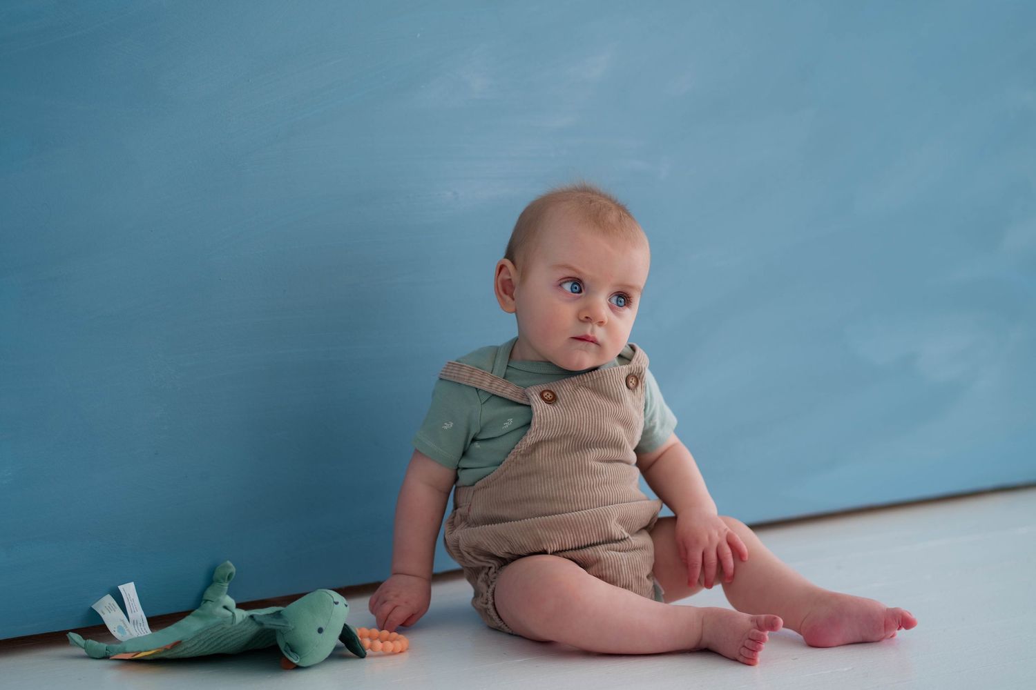 Baby boy in olive romper sitting on white floor with green toy duck against blue background.