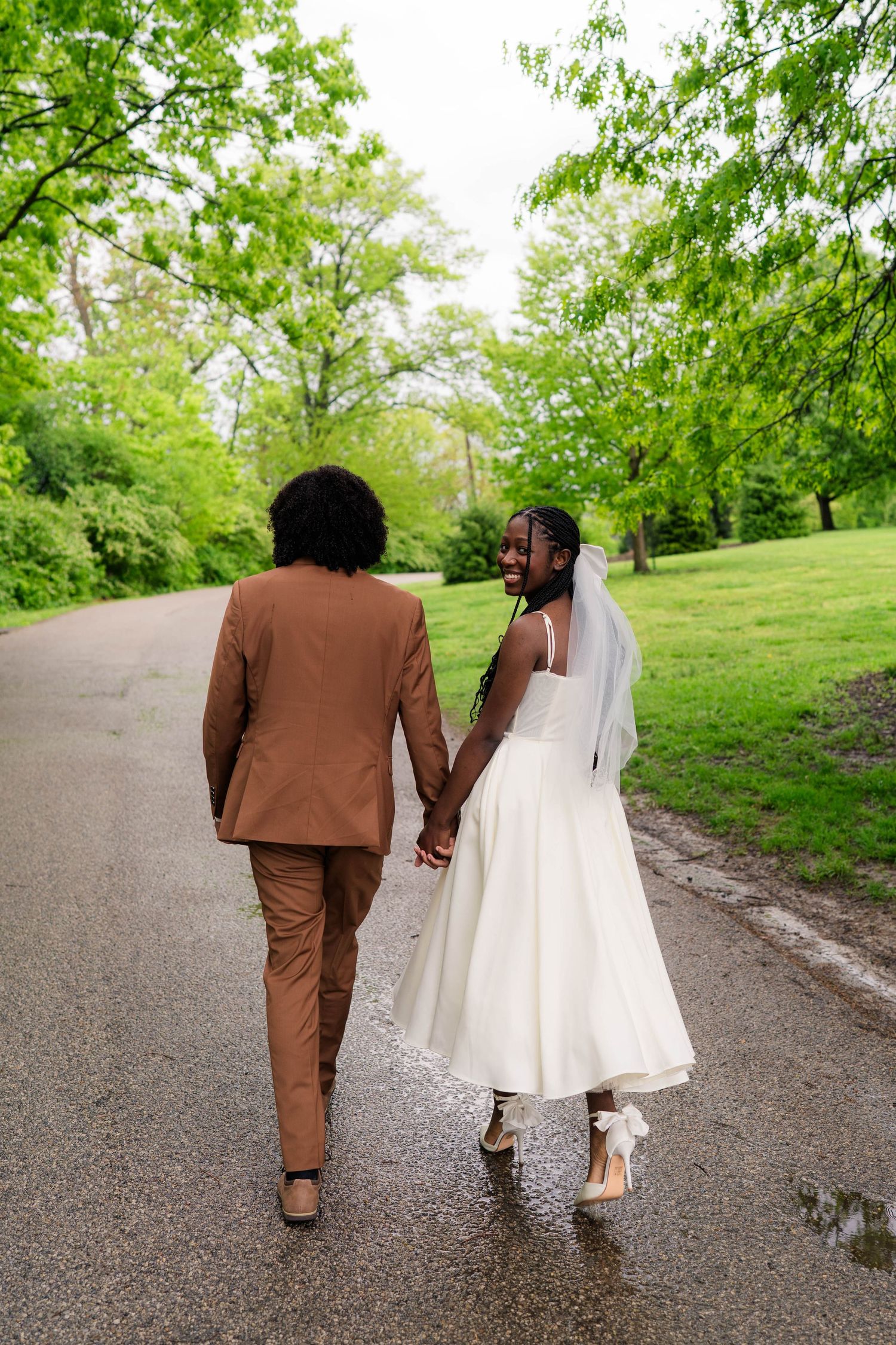 Bride looks back smiling as couple walks hand-in-hand down rain-wet park pathway.