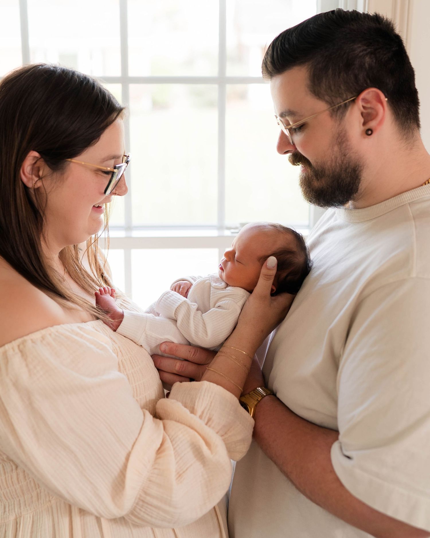 New parents lovingly gaze at their newborn baby, mom in white dress and dad in beige shirt, by a bright window.