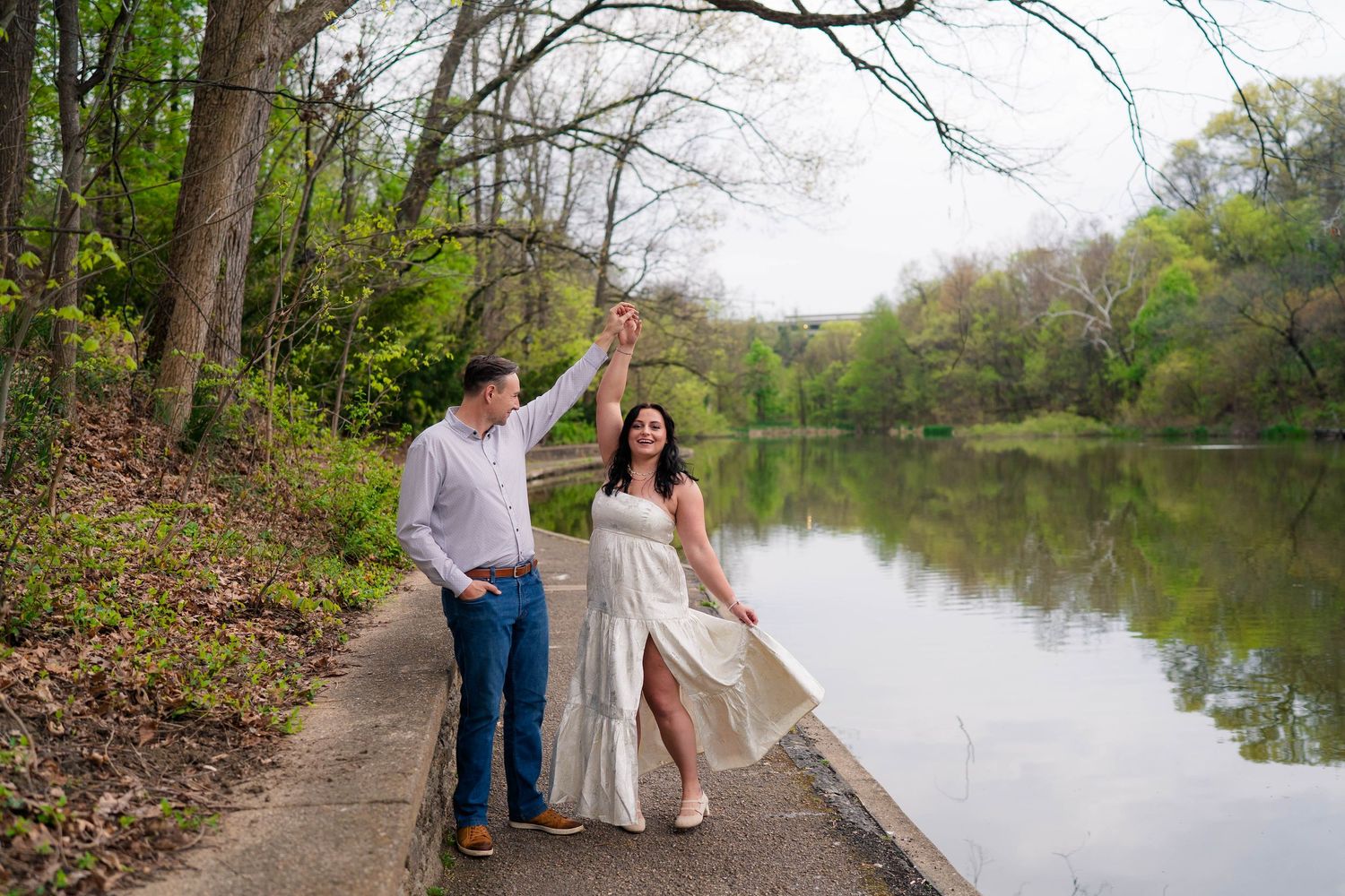 Couple dancing and holding hands along a lakeside path with vibrant green trees in the background.