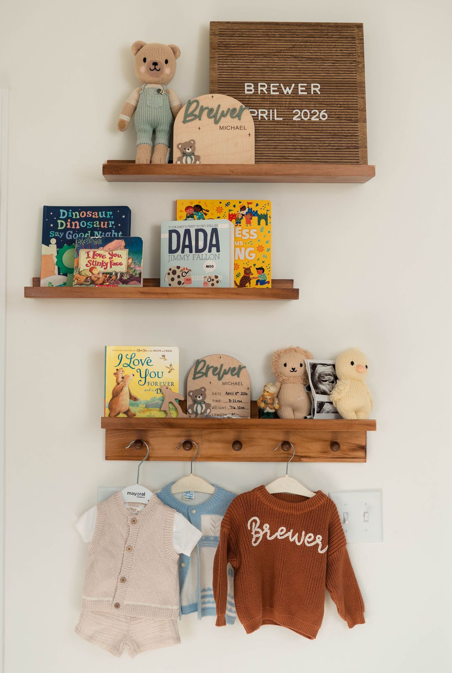 Nursery shelves displaying baby name Brewer, childrens books, stuffed animals, and hanging newborn outfits.