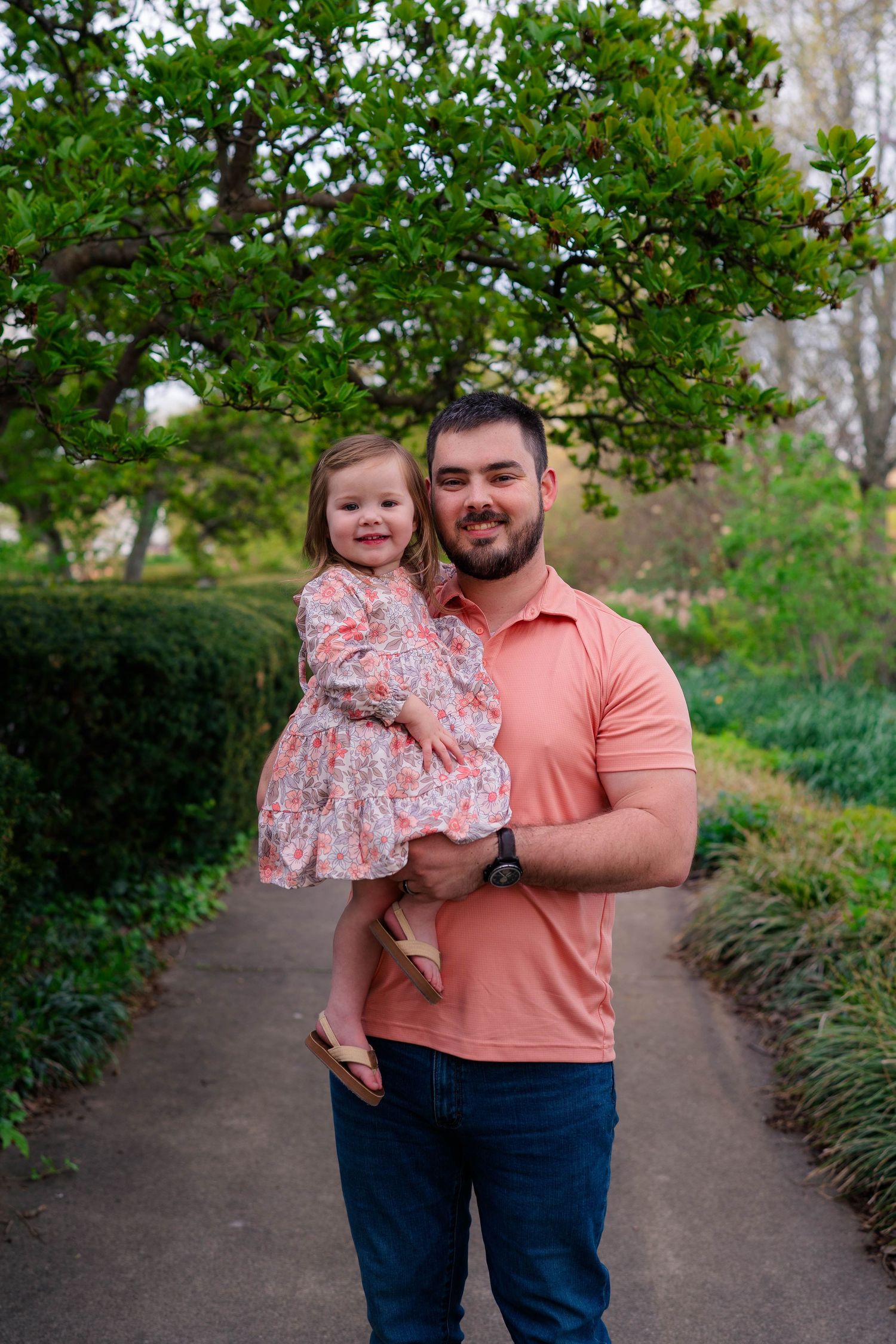 A smiling father in a salmon polo shirt holds his young daughter in a floral dress in a green park pathway.