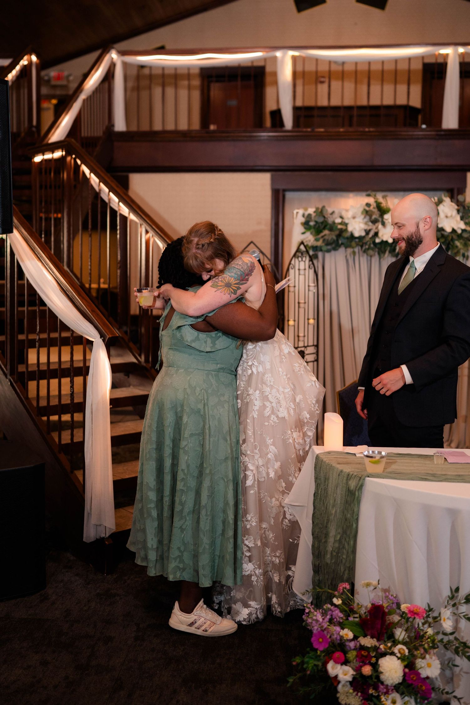 Bride and bridesmaid share emotional hug at wedding reception table while groom watches warmly nearby.