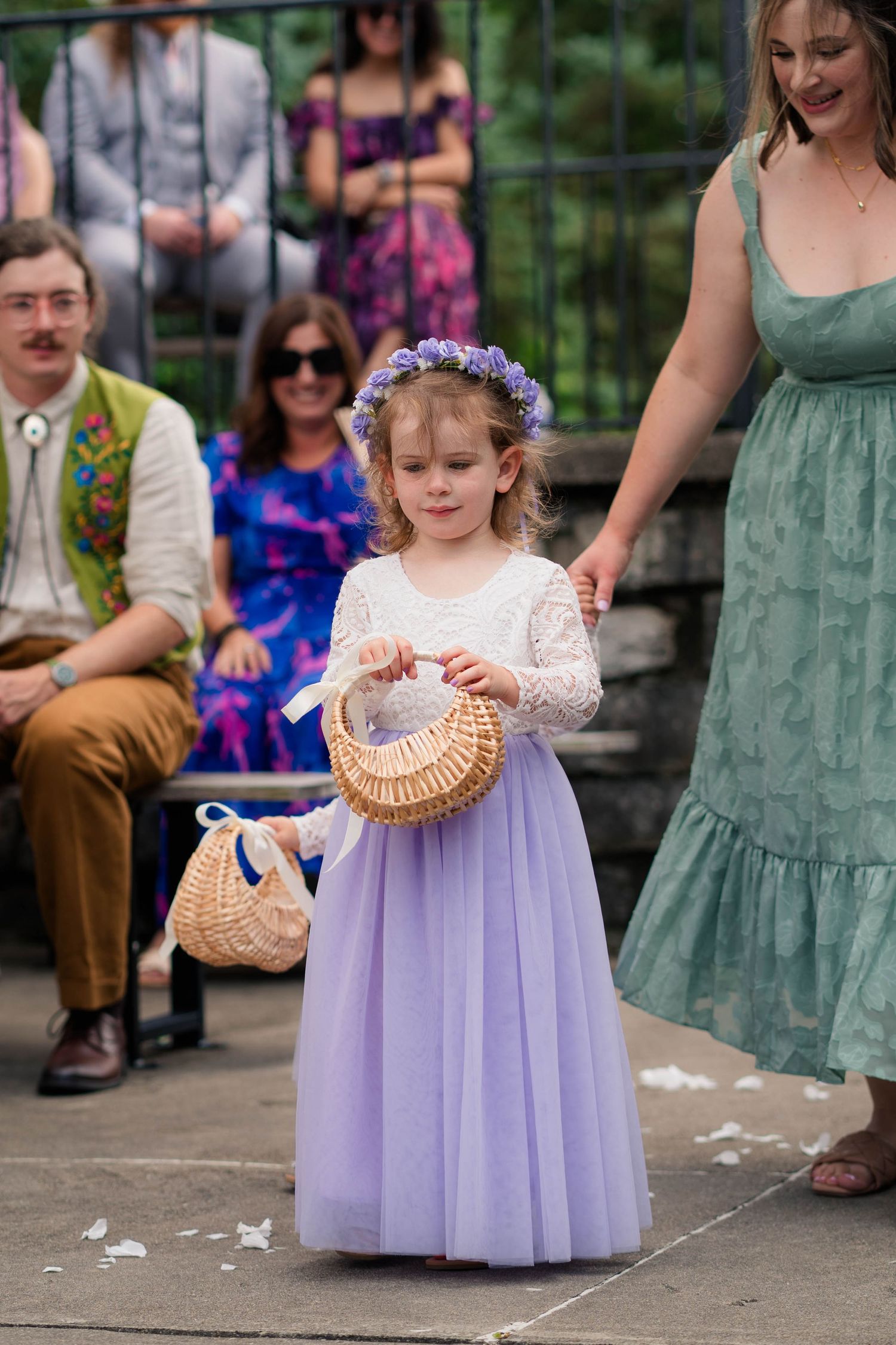Adorable flower girl in lavender tulle dress and purple floral crown carries wicker basket at outdoor wedding ceremony.