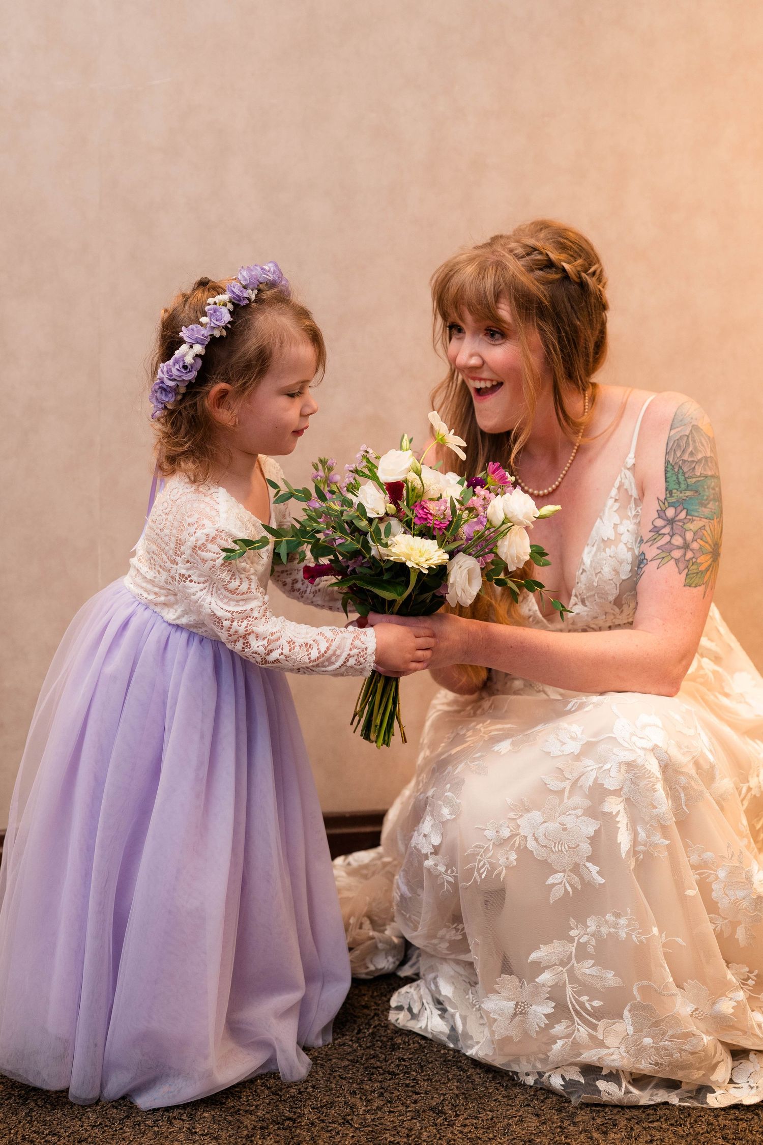 A bride in a floral lace gown laughs as a flower girl in purple presents her with a bouquet.