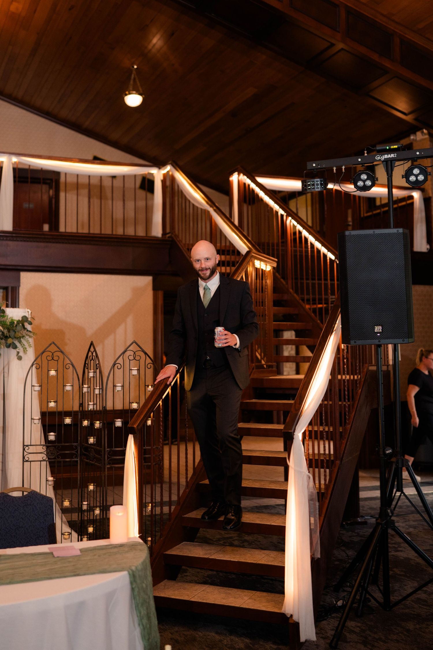 Bald groom in dark suit smiles while descending wooden staircase inside warmly lit wedding reception venue.