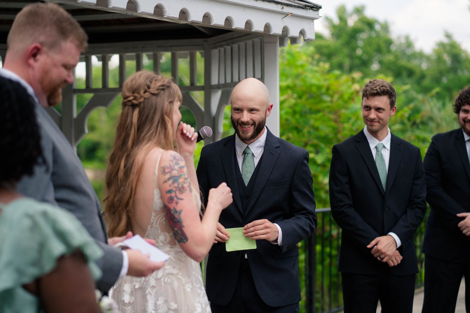 Bald groom smiles reading vows to tattooed bride during outdoor wedding ceremony with groomsmen nearby.