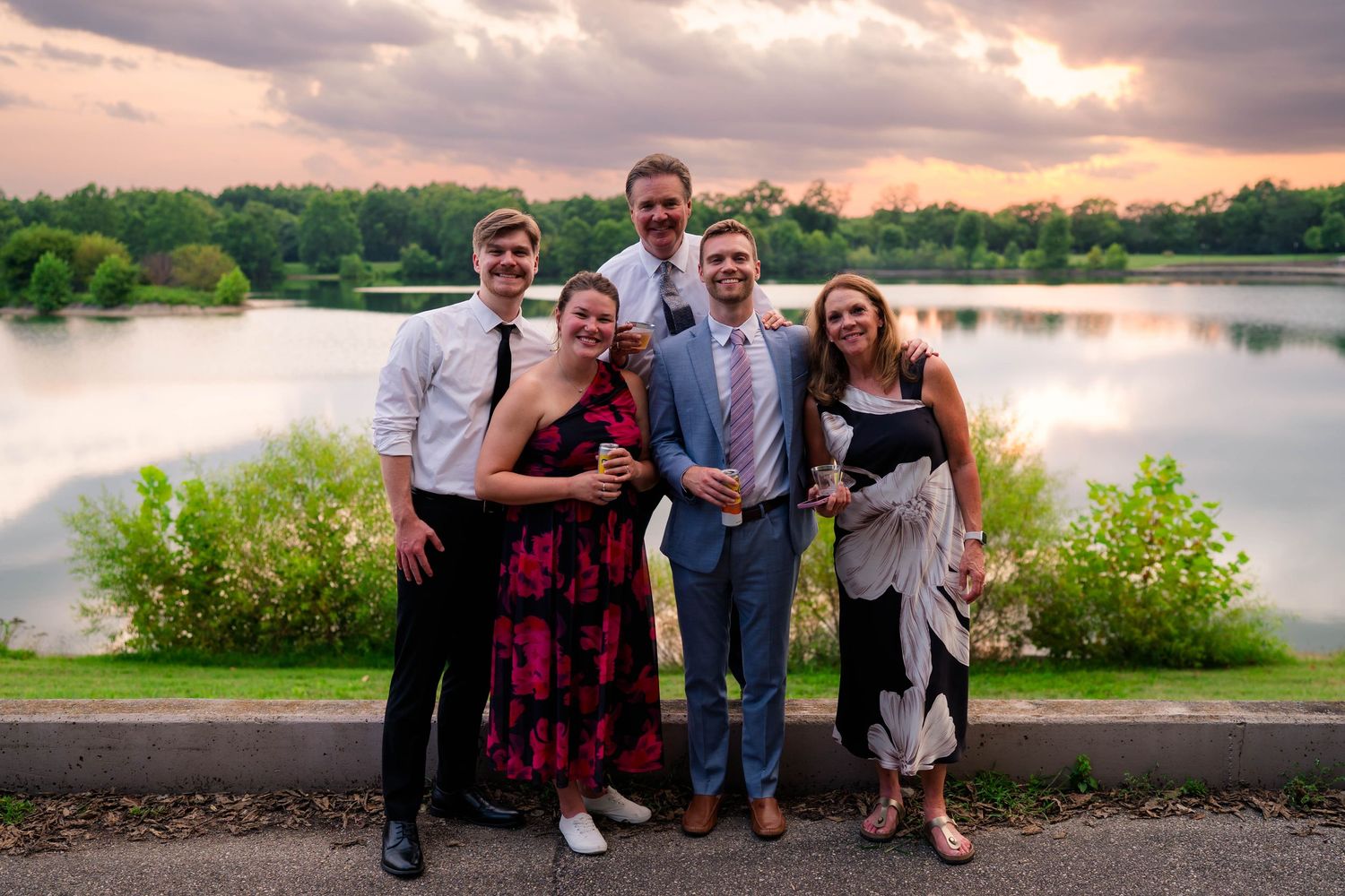 Group of five wedding guests posing outdoors by scenic lake at sunset.