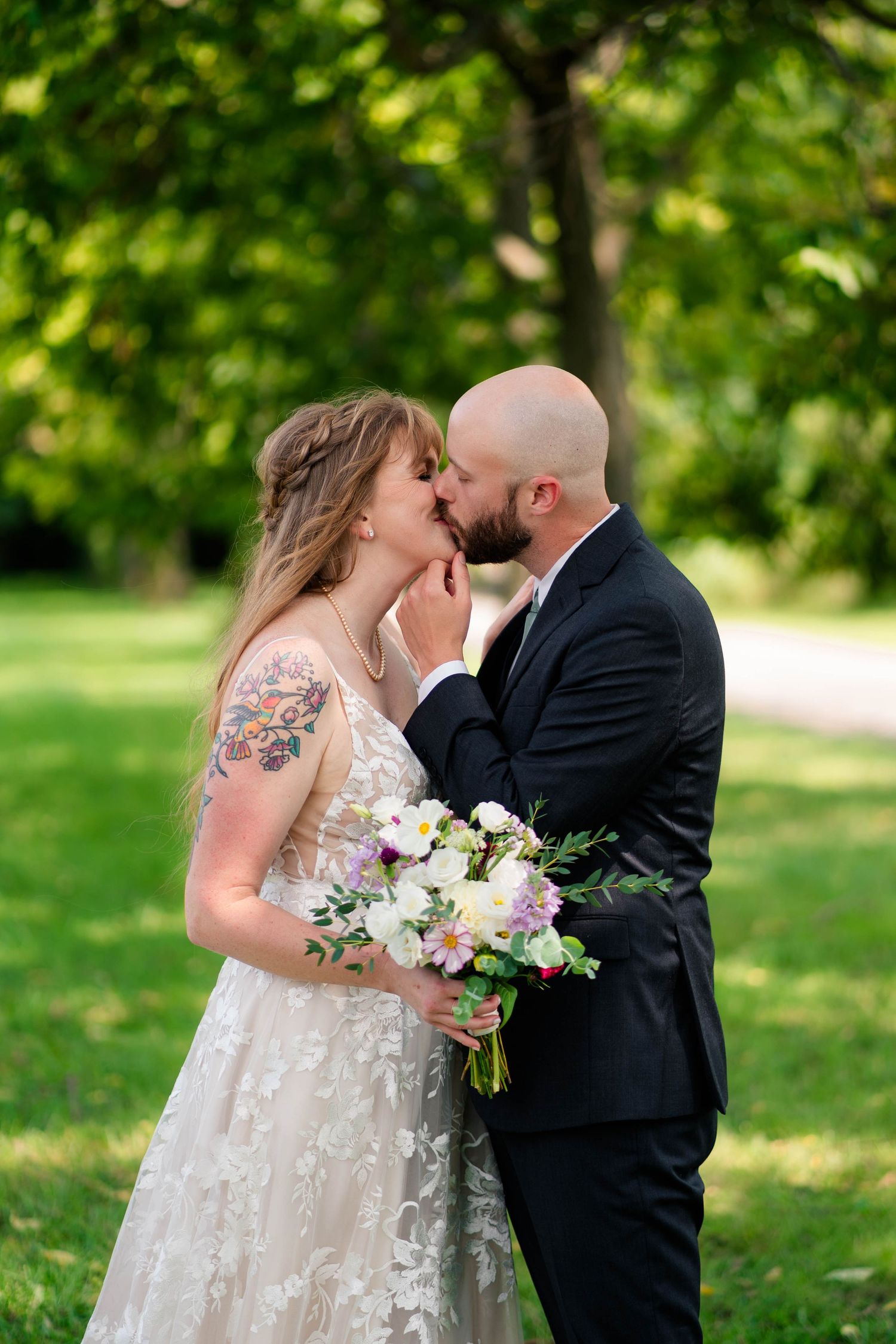 Bald groom tenderly kisses tattooed bride holding bouquet in sunlit green park.