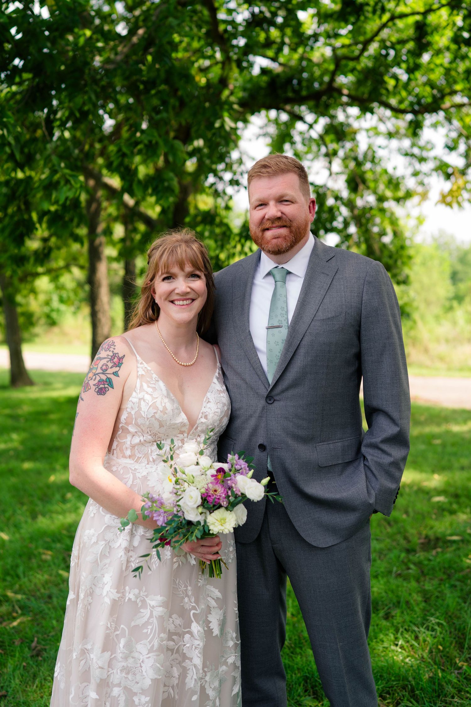 Smiling bride holding floral bouquet stands with bearded groom in gray suit outdoors under green trees at wedding.