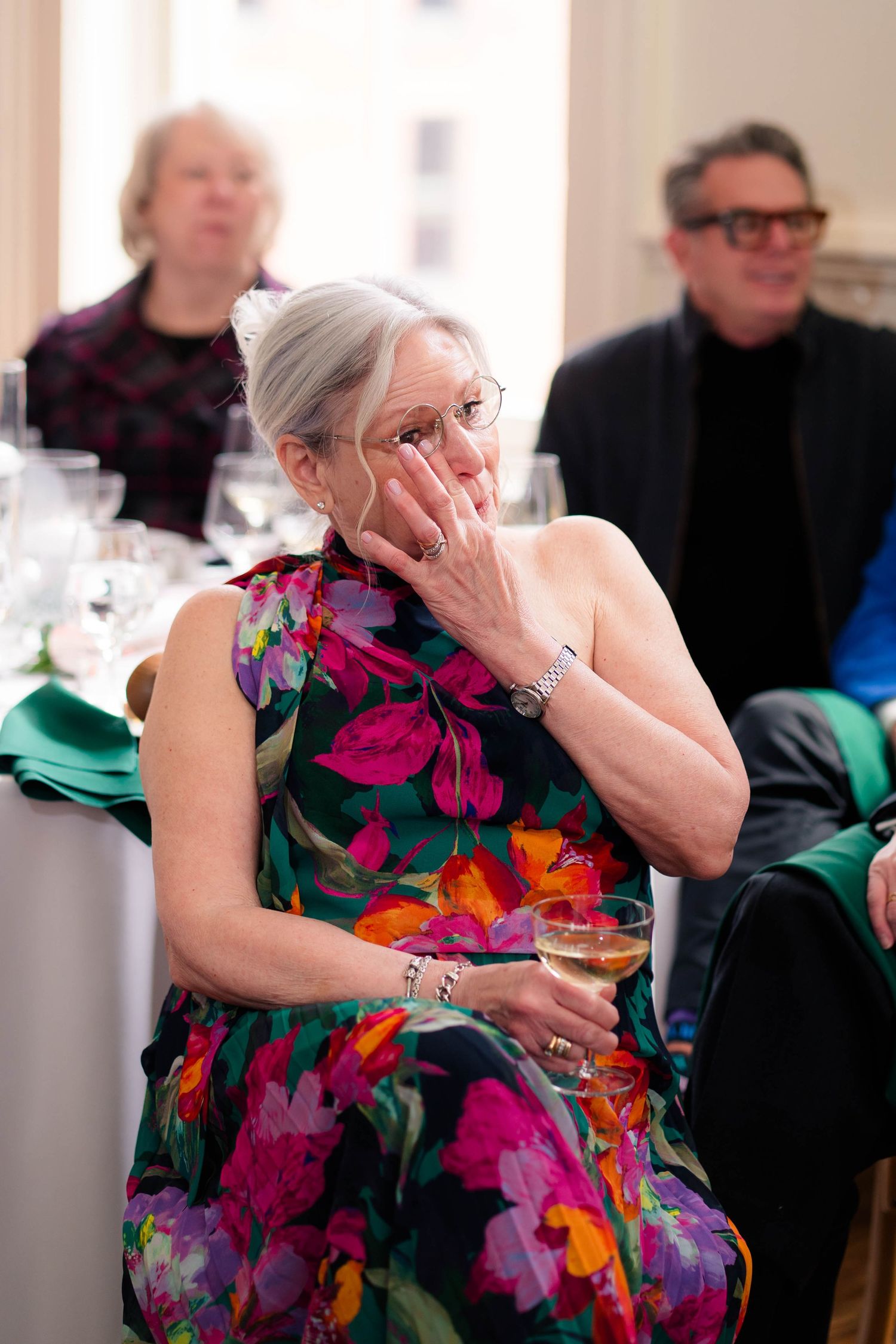 Emotional wedding guest in floral dress wipes tear while holding champagne glass.