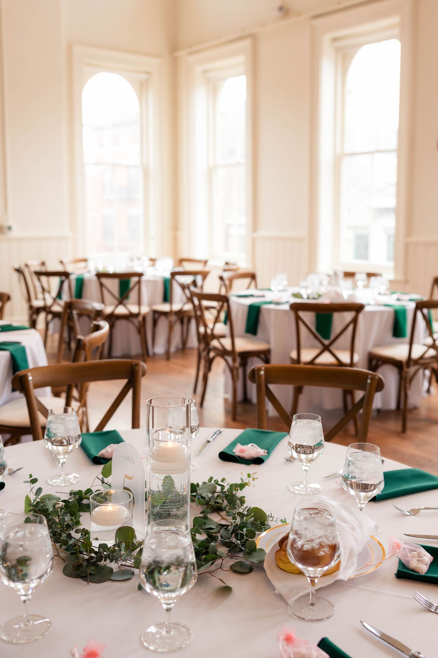 Wedding reception tables elegantly set with emerald napkins, greenery centerpieces, and floating candles in tall glasses.