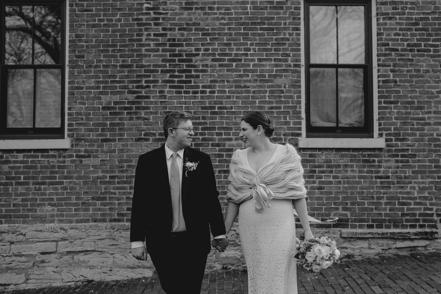 Black and white photo of a bride and groom holding hands and smiling at each other in front of a brick wall.
