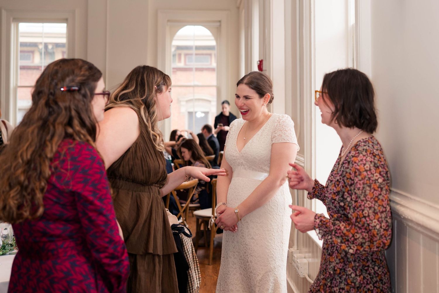Bride in white lace gown laughing with three guests at wedding reception.