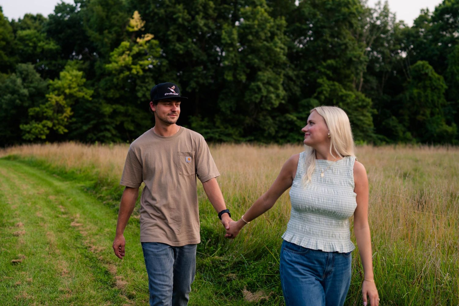 Couple holding hands walking along a grassy path with tall golden grass and lush green trees in background.