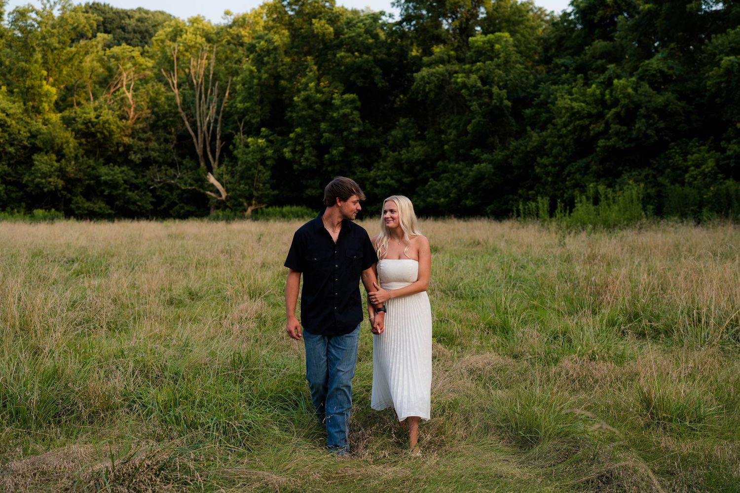 Happy couple holding hands and walking through a scenic green meadow at golden hour sunset.