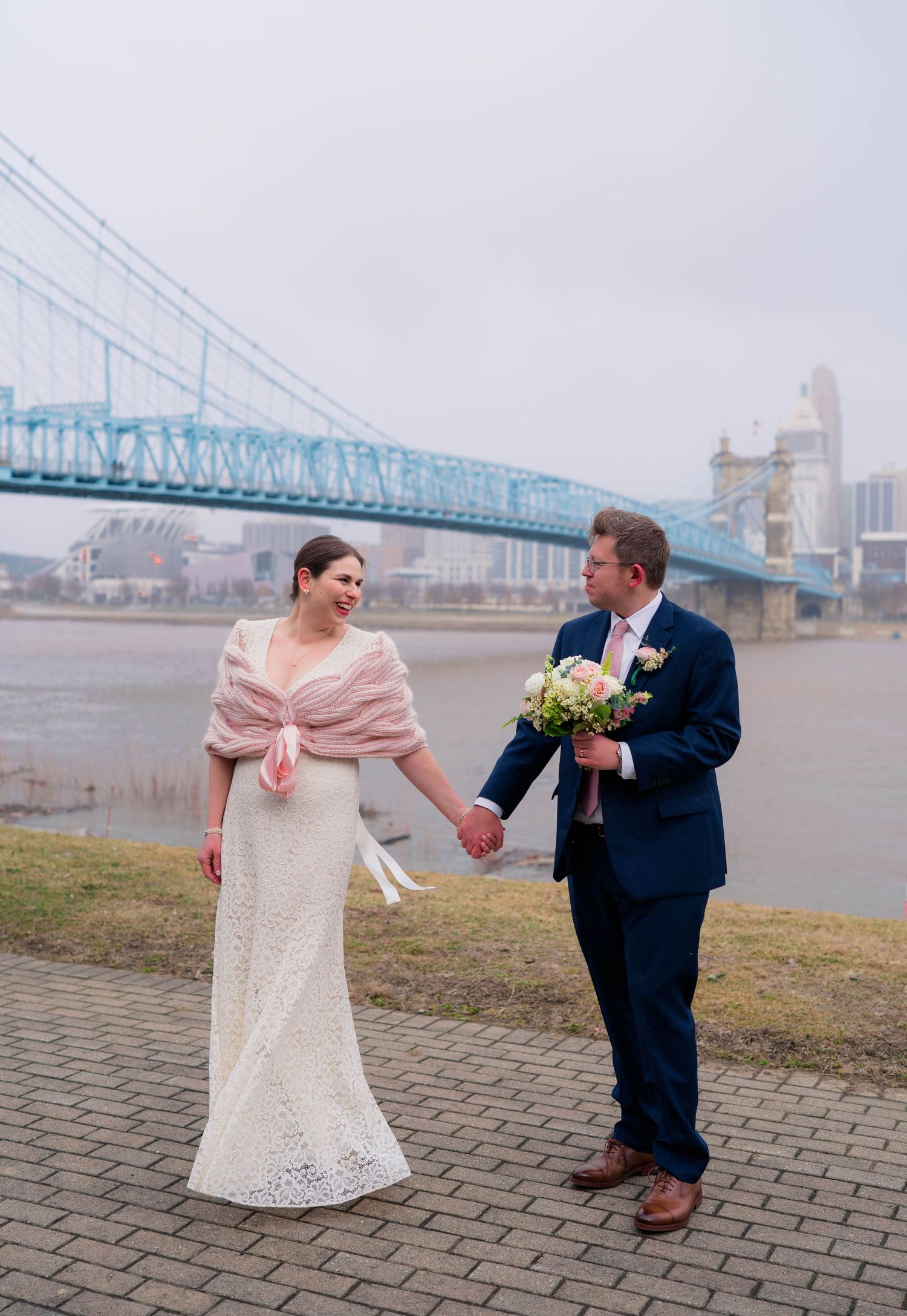 Bride and groom holding hands and walking along a riverfront path with a blue suspension bridge behind them.