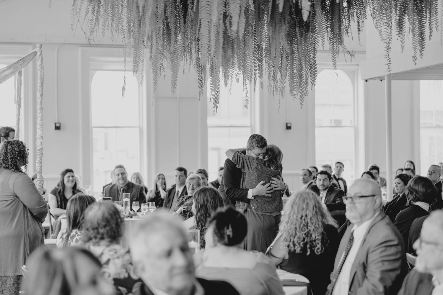 Black and white photo of couple embracing on dance floor surrounded by wedding reception guests.