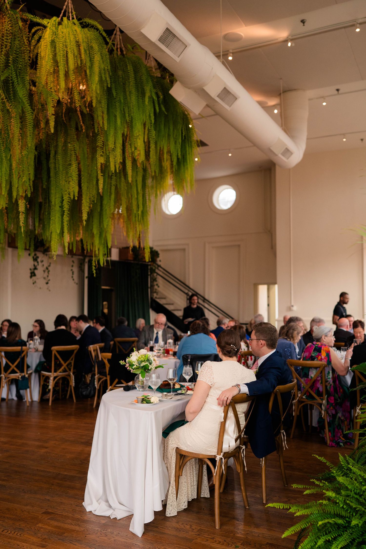Bride and groom sit at sweetheart table beneath lush hanging ferns at wedding reception.