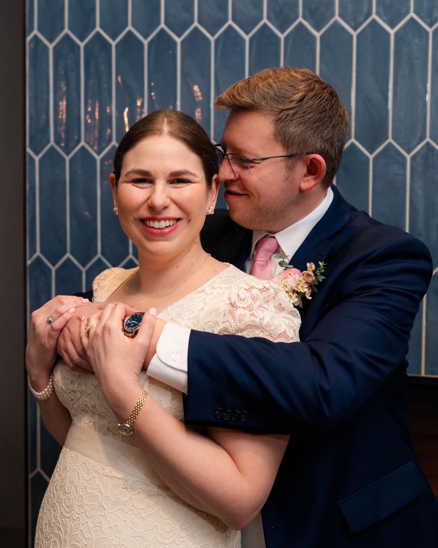 Smiling bride and groom embracing joyfully in front of a blue picket tile wall at their wedding venue.