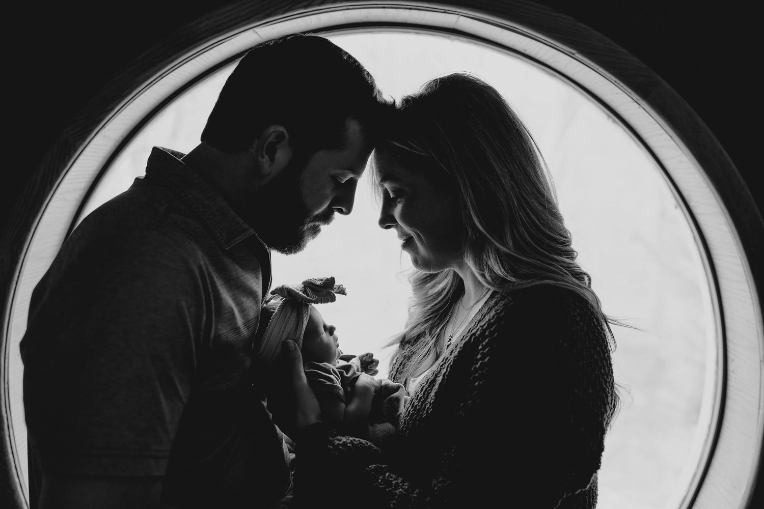 Black and white silhouette of new parents cradling their newborn in front of a circular window.